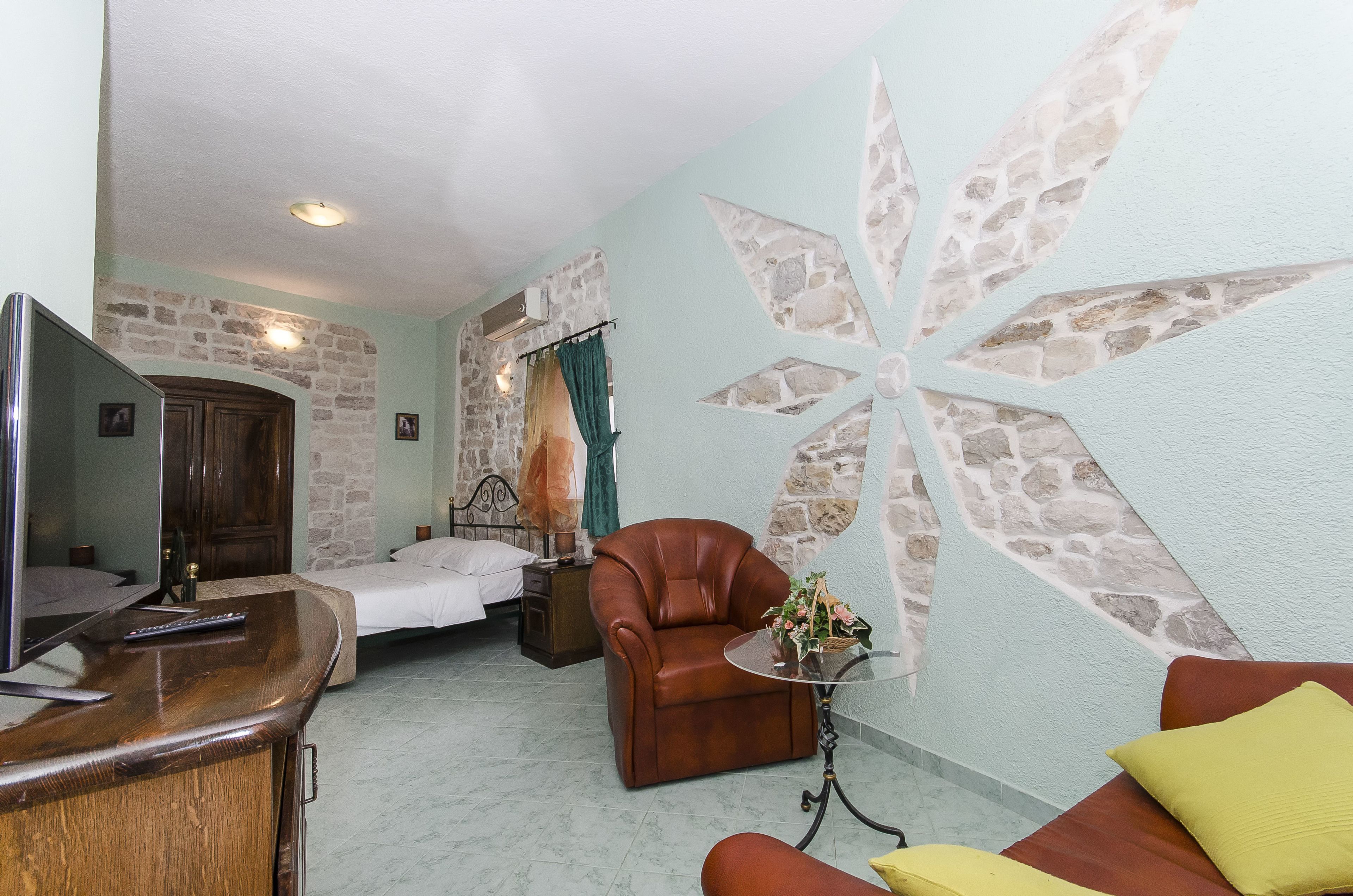 A hotel room with a bed, a TV, a leather armchair, a small table with a vase, and a stone wall with a geometric design.