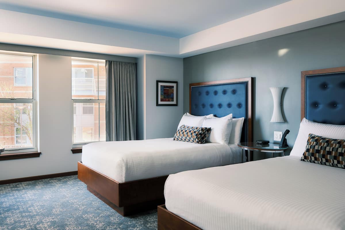 A hotel room with two beds, a large window, and a view of buildings outside.