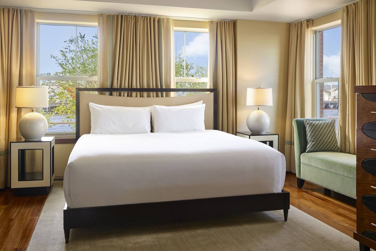 A hotel room with a large bed, two bedside tables with lamps, a green armchair, and a view of the outdoors through large windows.