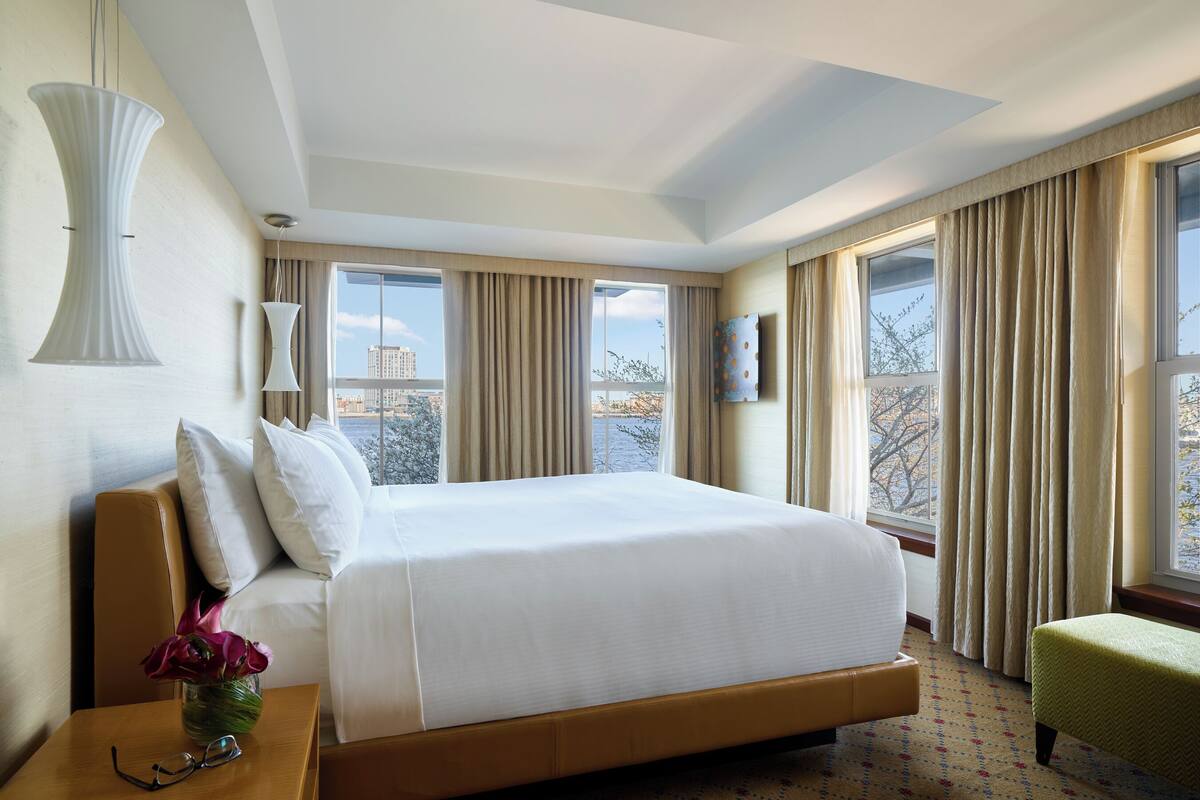 A hotel room with a large bed, a nightstand with a vase of flowers, a green armchair, and a view of the city through large windows.