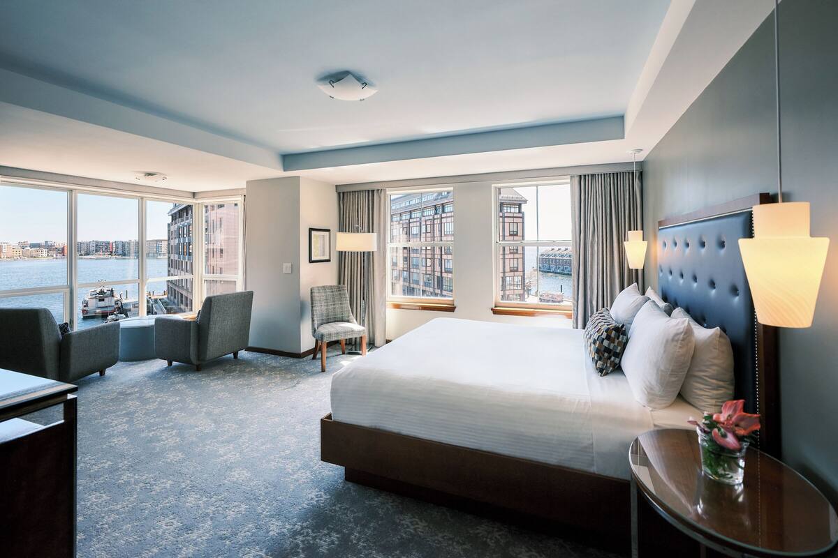 A modern hotel room with a large bed, a seating area with a chair, a small table with a vase, and large windows offering a view of the city and water.
