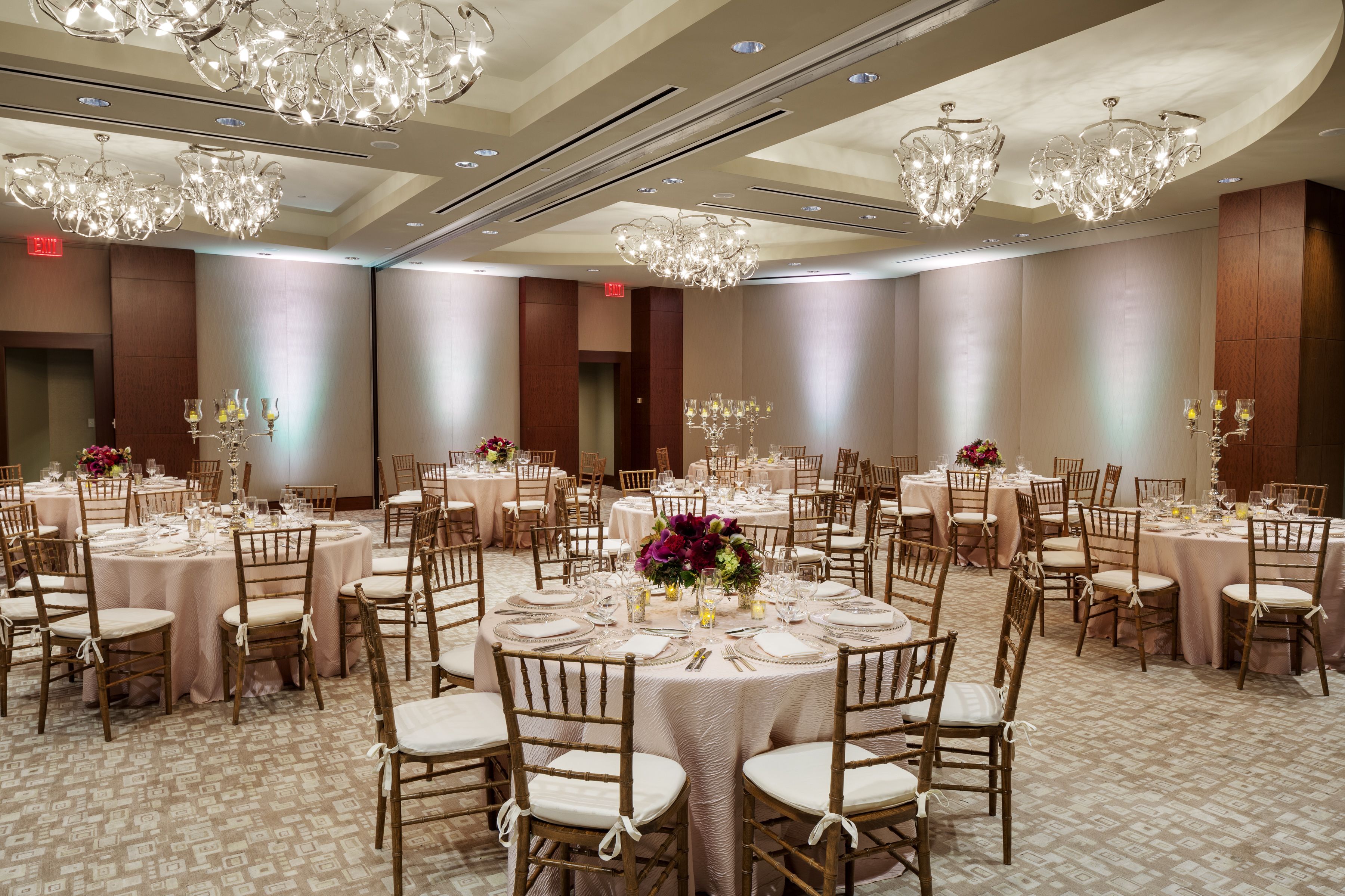 A banquet hall with round tables, wooden chairs, and chandeliers.