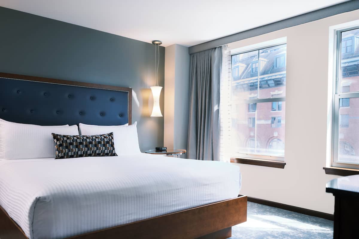 A hotel room with a large bed, a headboard, a bedside table, a lamp, and a window with a view of buildings.