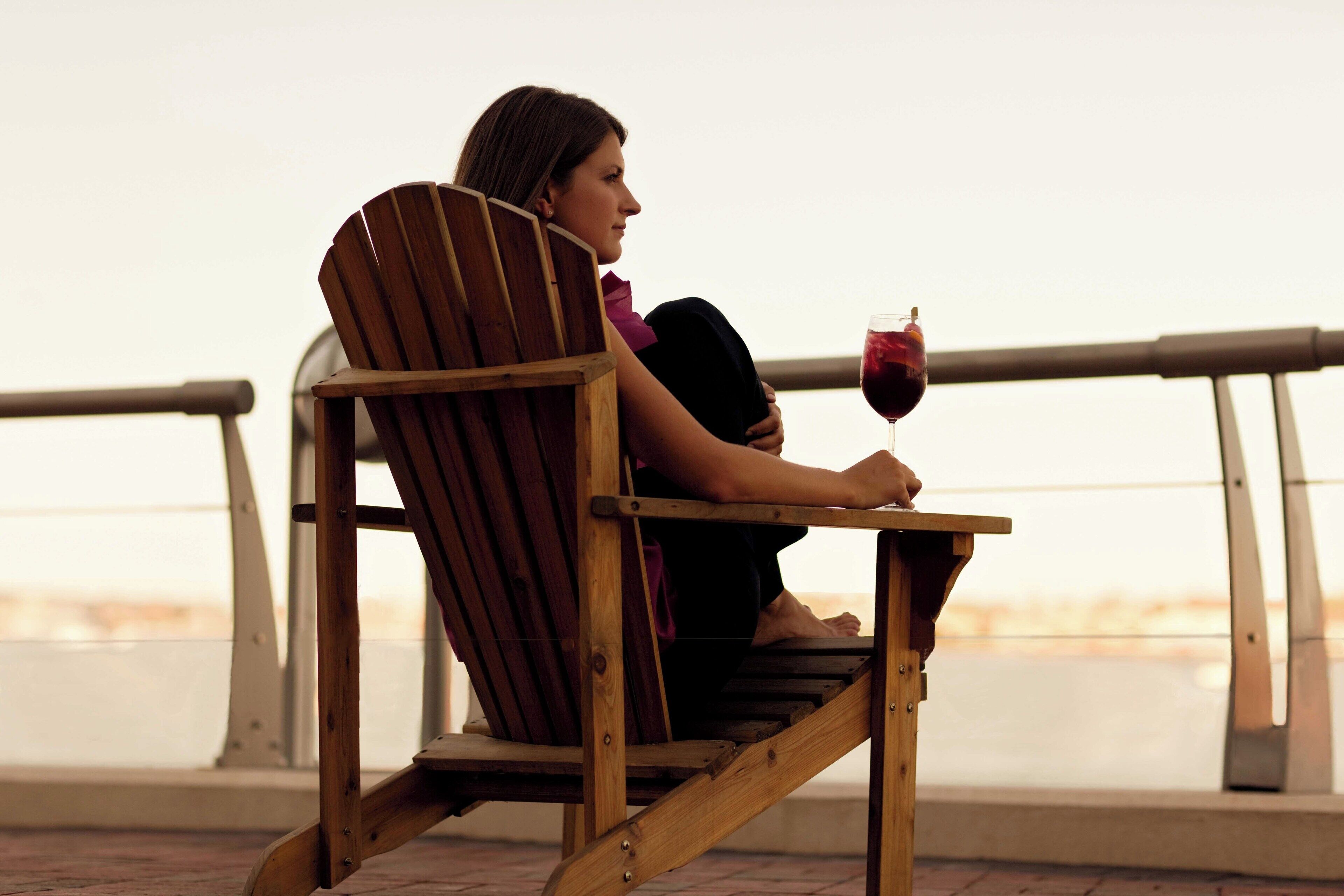 A person seated on a wooden chair with a glass of red wine, overlooking a body of water.