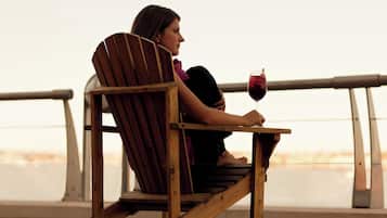 A person seated on a wooden chair with a glass of red wine, overlooking a body of water.