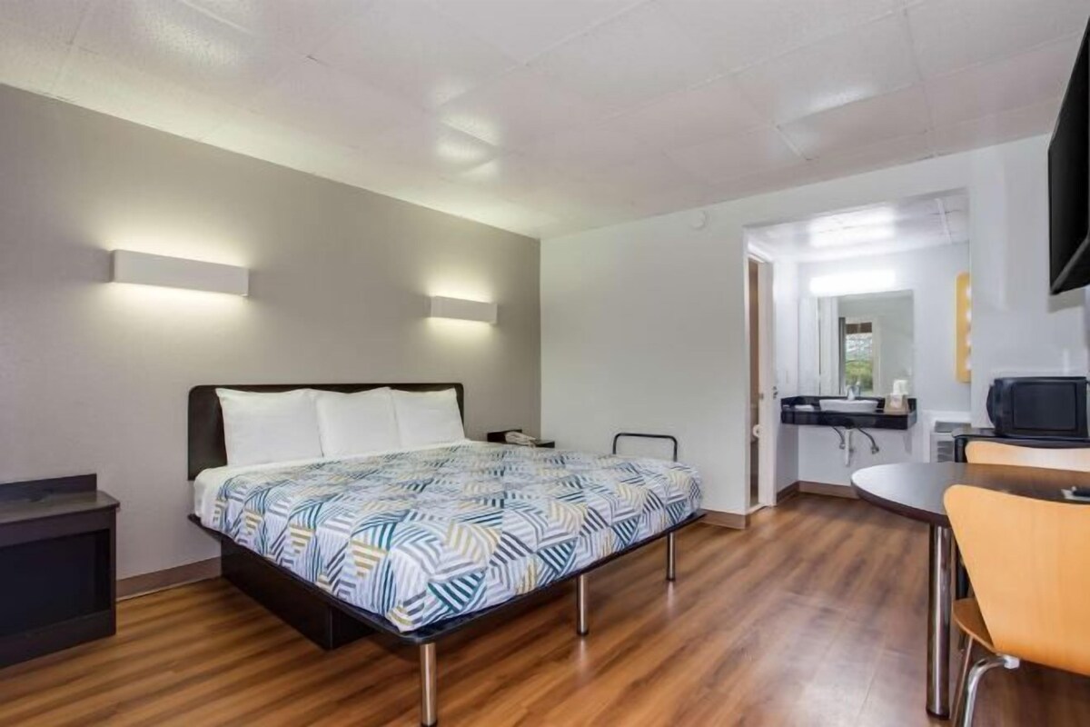 A modern hotel room with a bed, bedside table, and a small kitchenette area.
