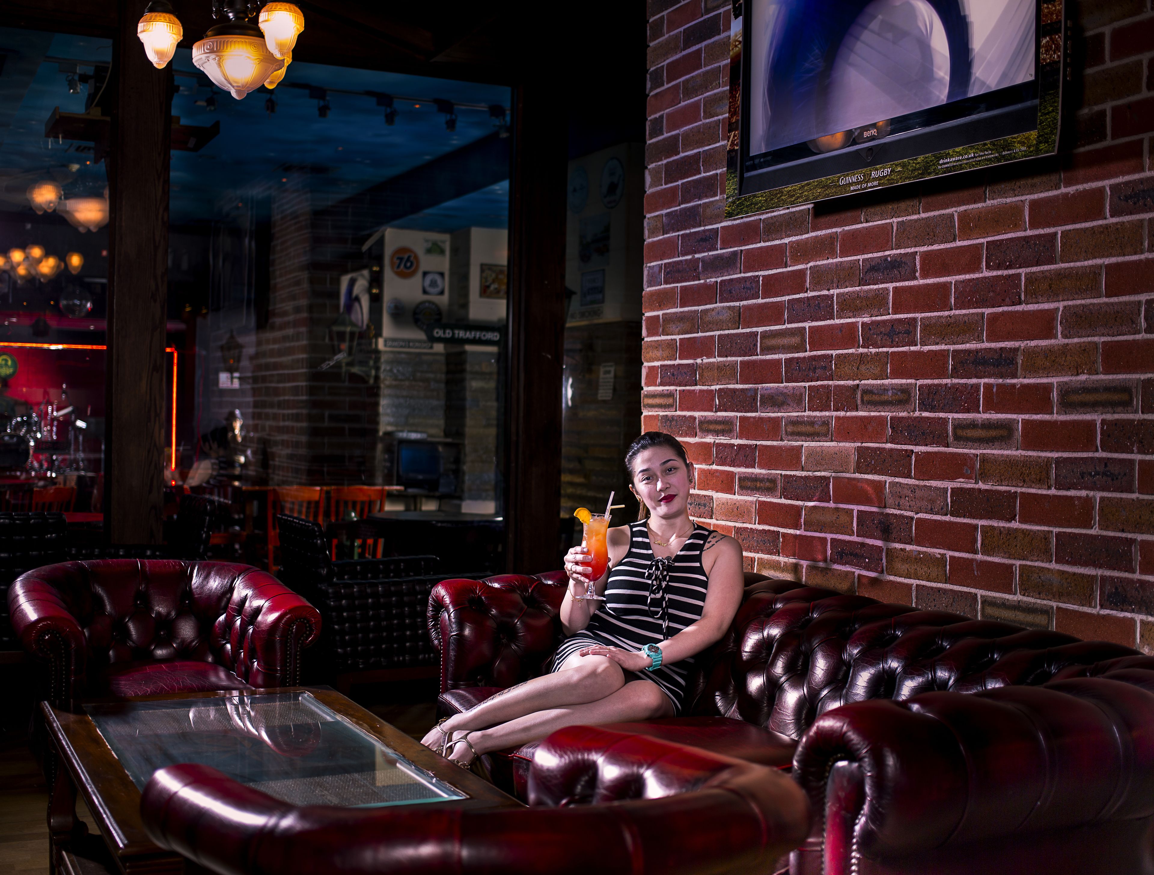 A woman sitting on a red leather couch in a bar with a television mounted on the brick wall.
