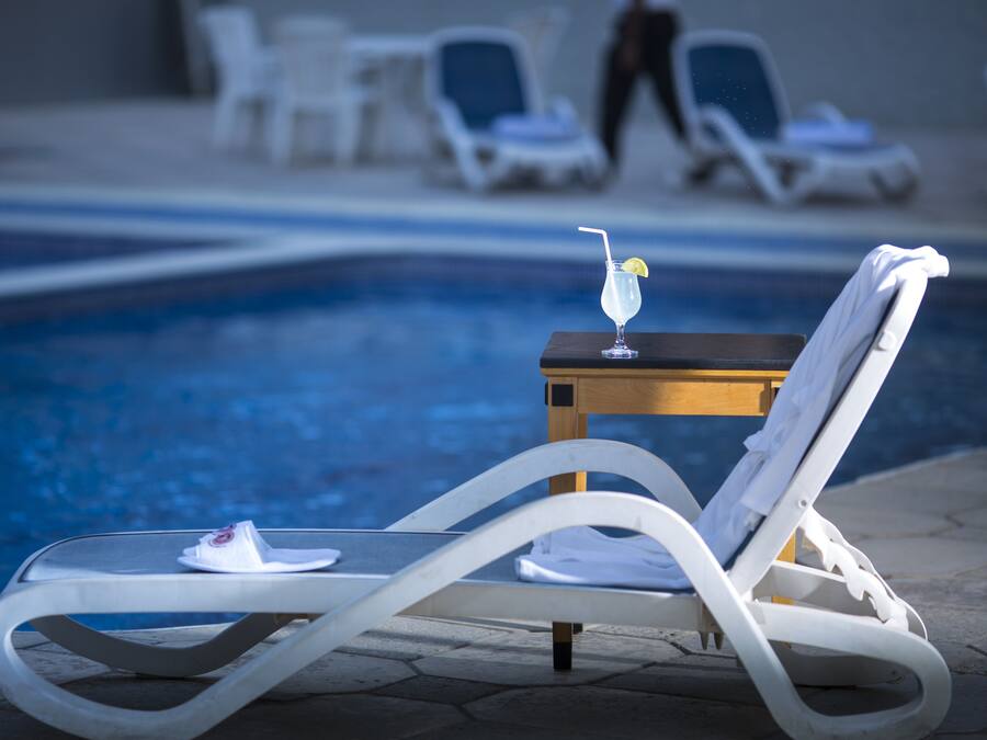 Ein Poolbereich mit Liegestühlen, einem kleinen Tisch mit einem Getränk und einer Person, die im Hintergrund geht.
