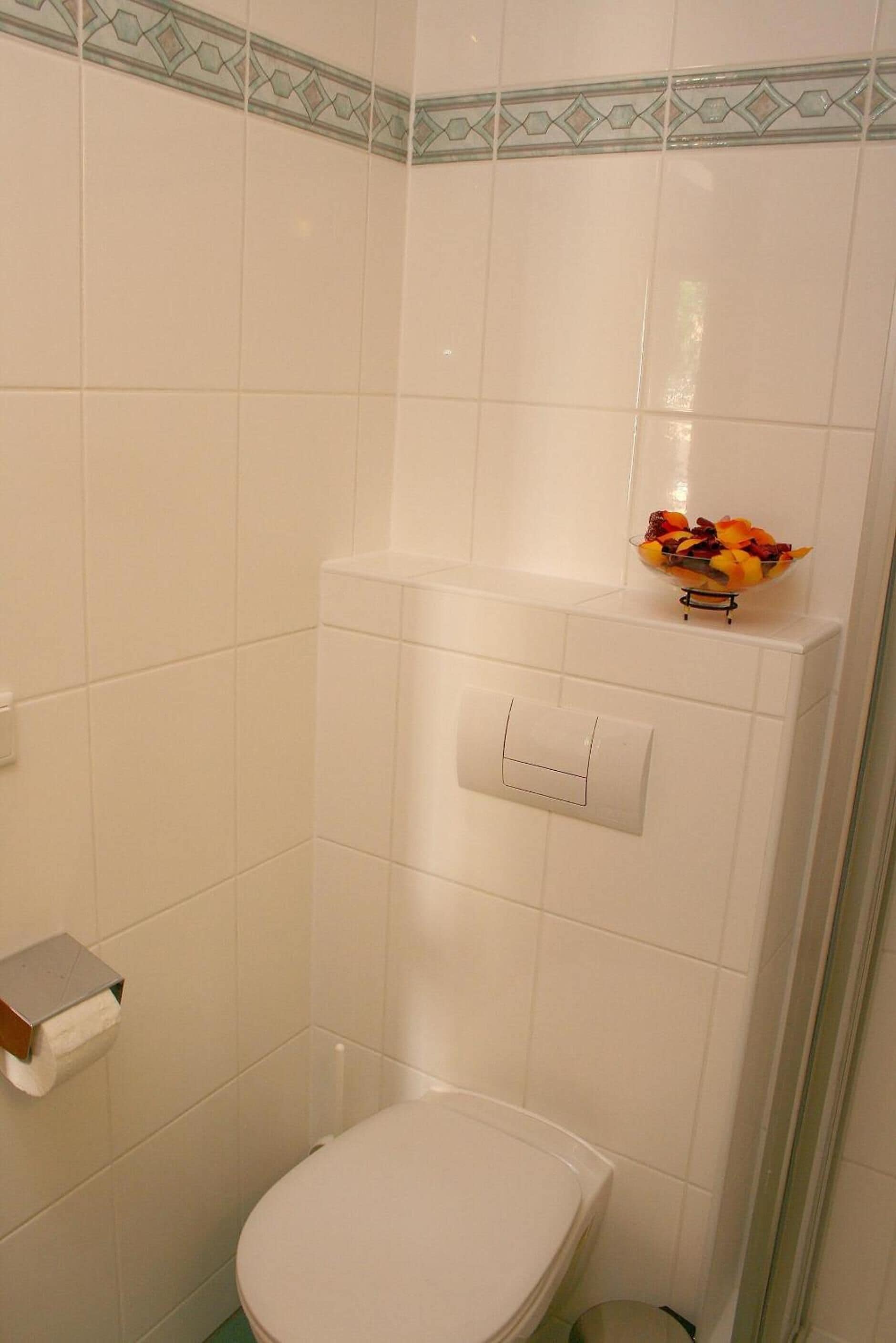 A bathroom with white tiles, a toilet, a small shelf with a fruit bowl, and a paper towel holder.