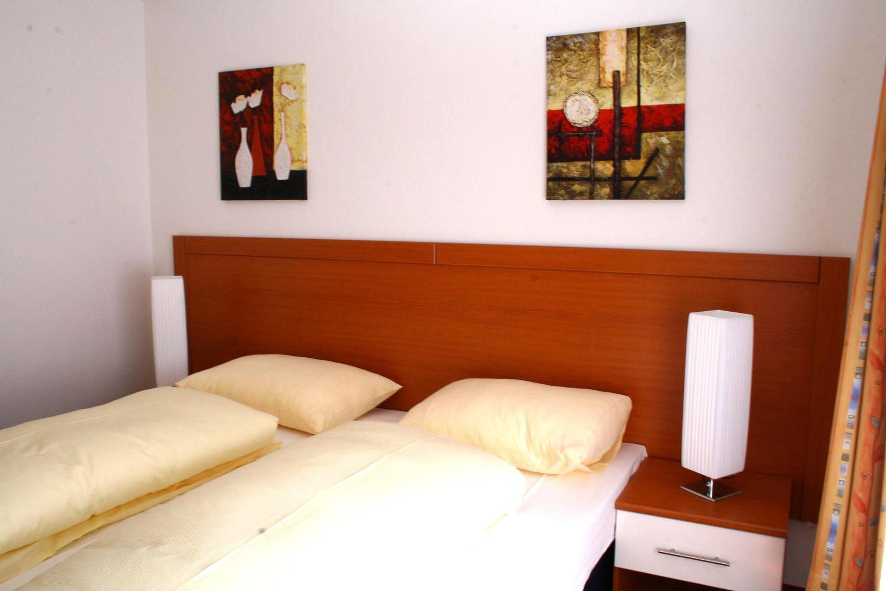 Un lit double avec deux oreillers, une tête de lit en bois, une table de chevet avec une lampe et deux œuvres d’art accrochées au mur.