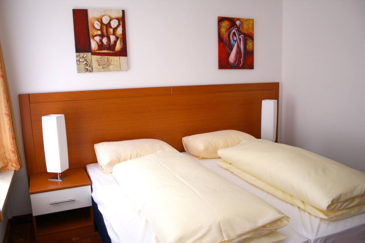 A hotel room with a wooden headboard, two beds with white bedding, a bedside table with a lamp, and two pieces of wall art.