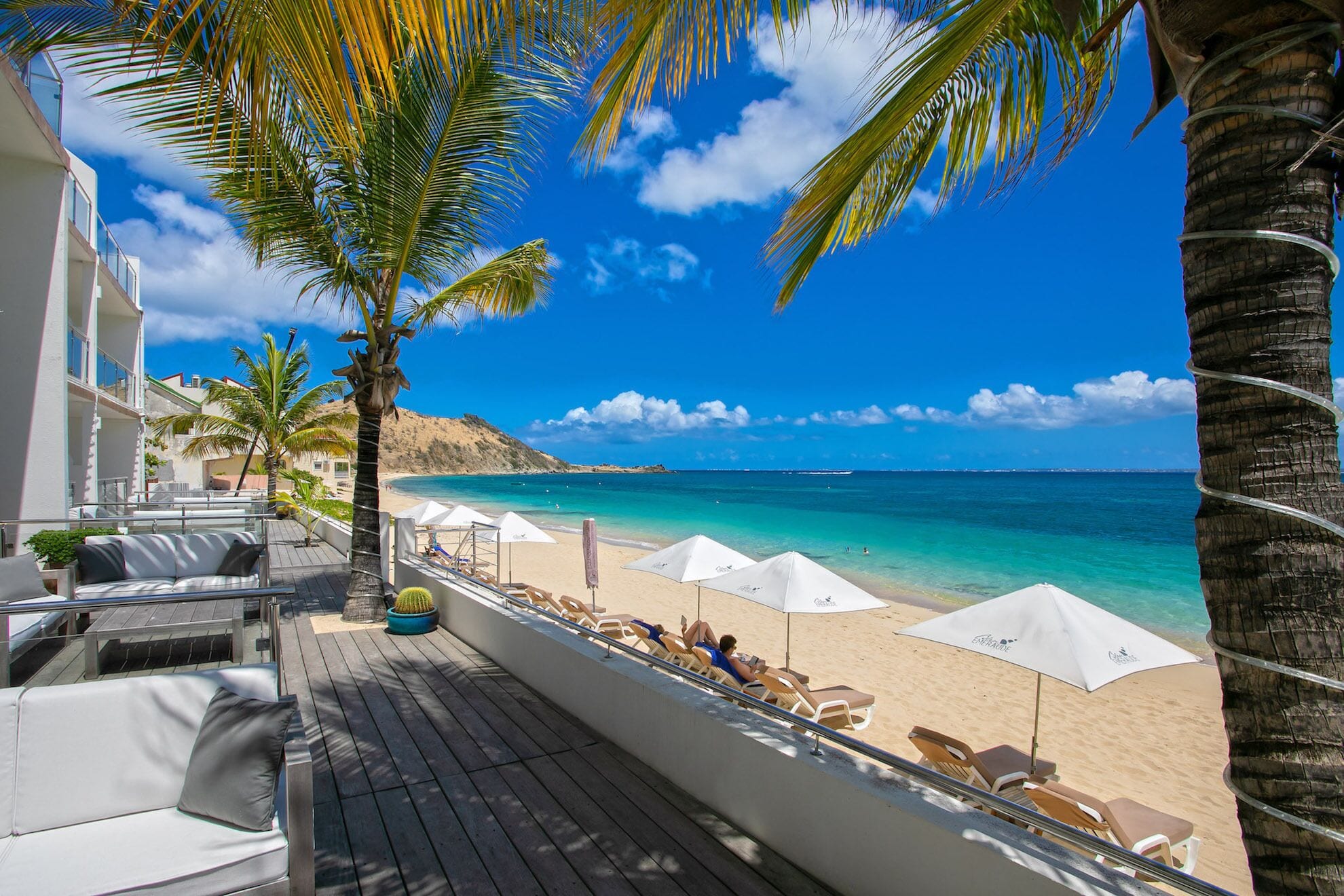 A beachside balcony with lounge chairs and umbrellas, overlooking a clear blue sea and a sandy beach.