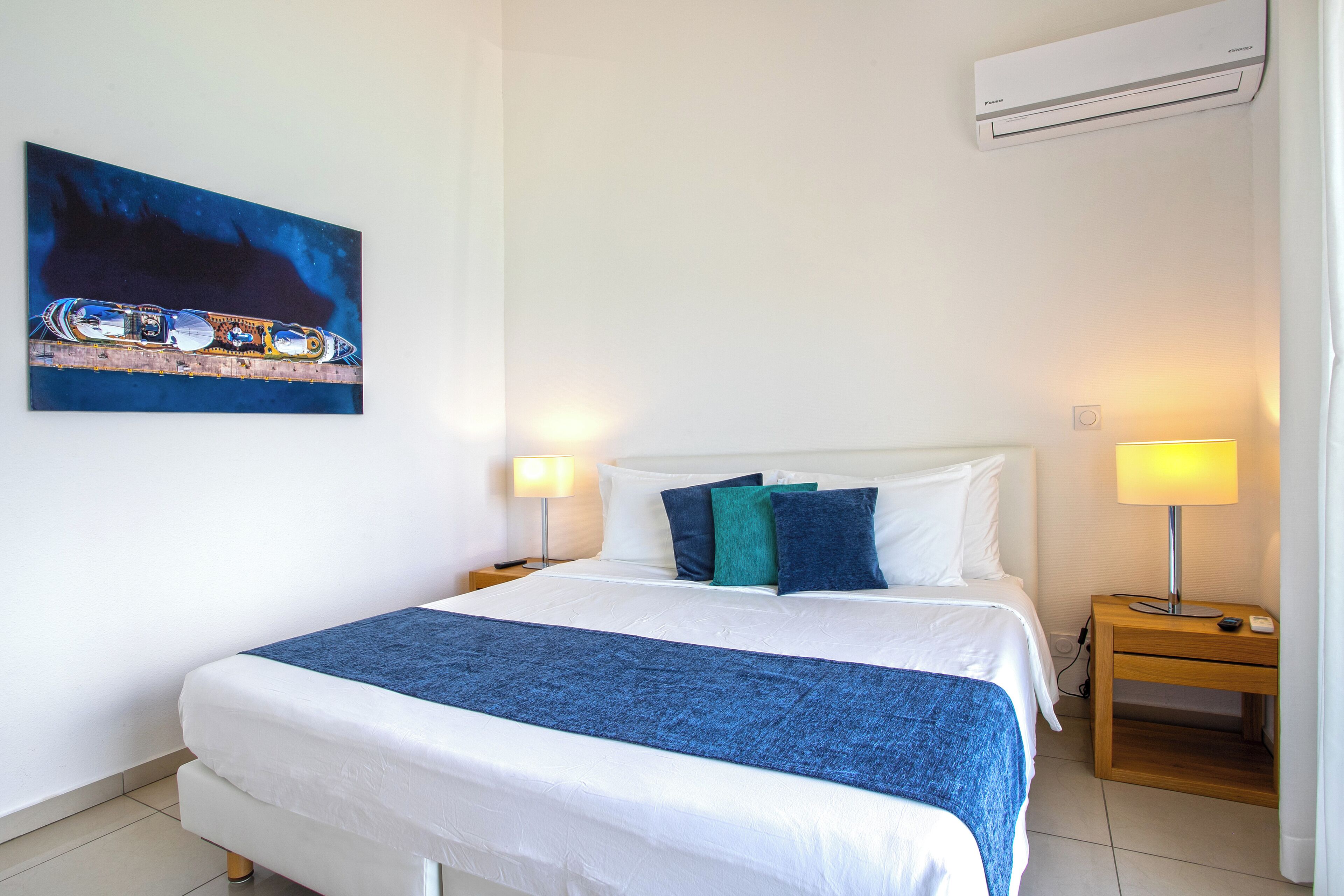 A neatly made bed with a blue blanket, two bedside lamps, and a wall-mounted air conditioner.