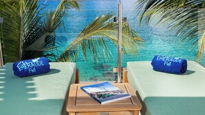 A balcony with two lounge chairs, each with a blue towel labeled "Emerald FWI". A wooden table with a book is in the center. - Bleu Emeraude Hotel (Grand Case)