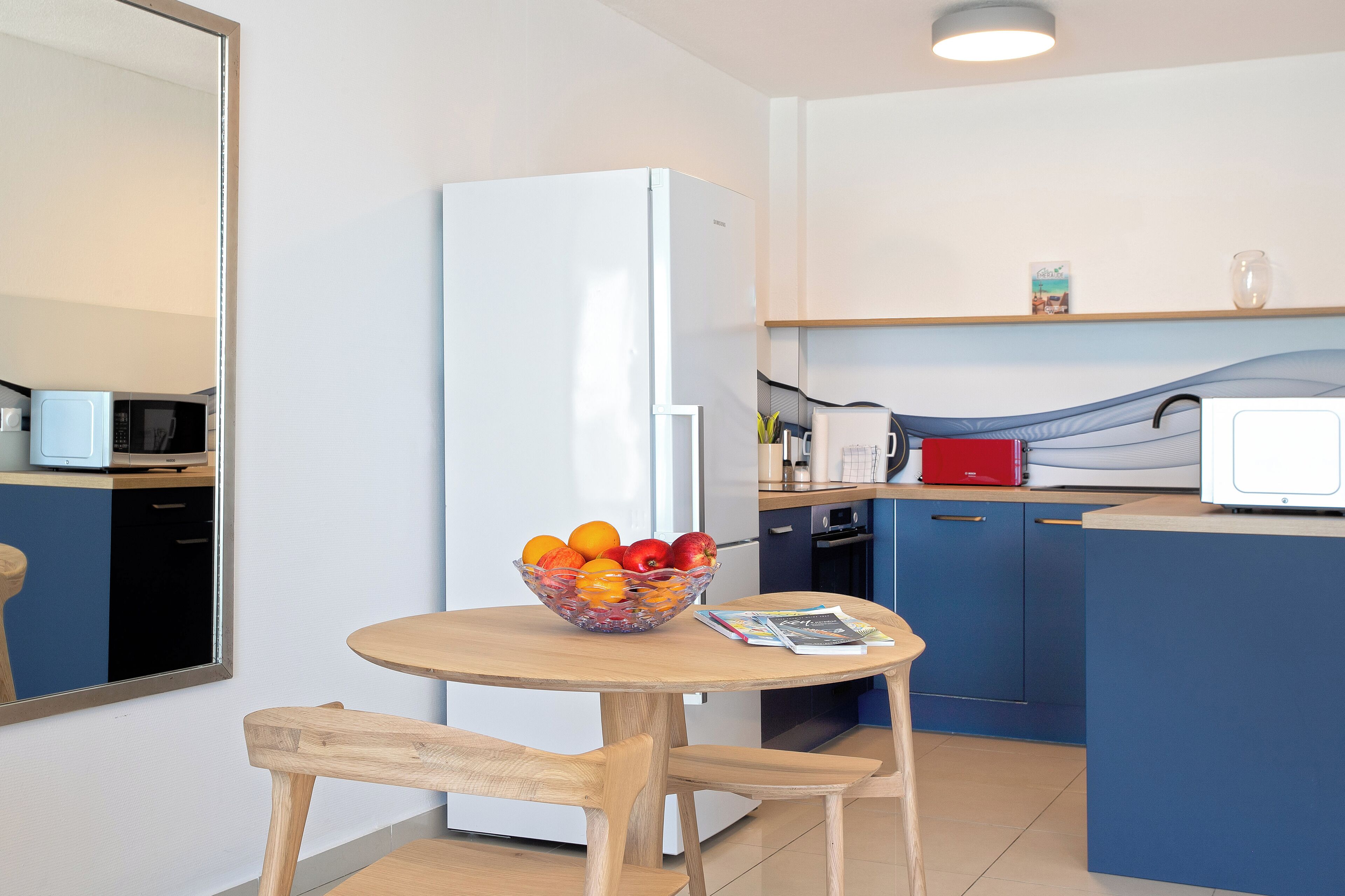 Een kleine keuken met een koelkast, magnetron en een fruitschaal op een houten tafel.