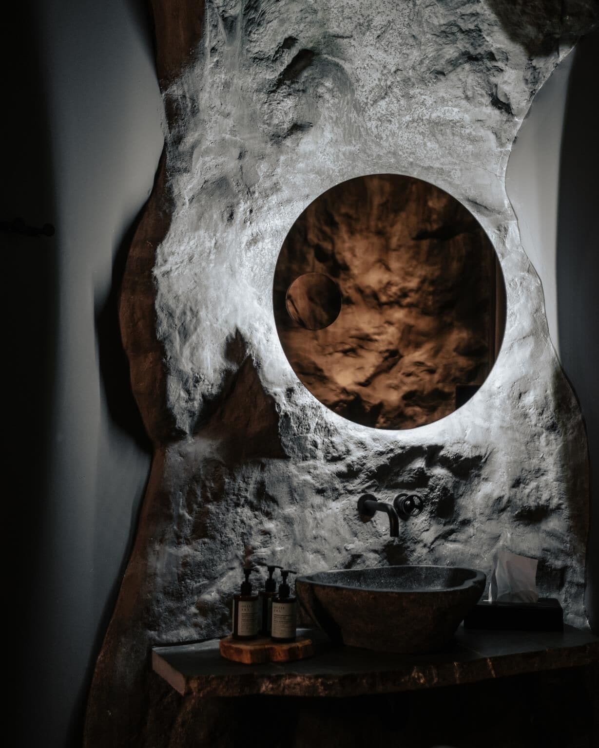 A bathroom with a stone sink, a wooden basin, and a circular mirror.