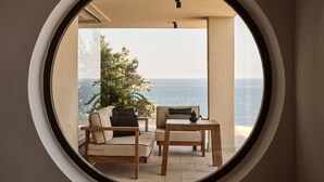 A circular window view of a balcony with a sofa and a table, overlooking a body of water and a tree. - Seaside A Lifestyle Resort All Inclusive (Malevizi)