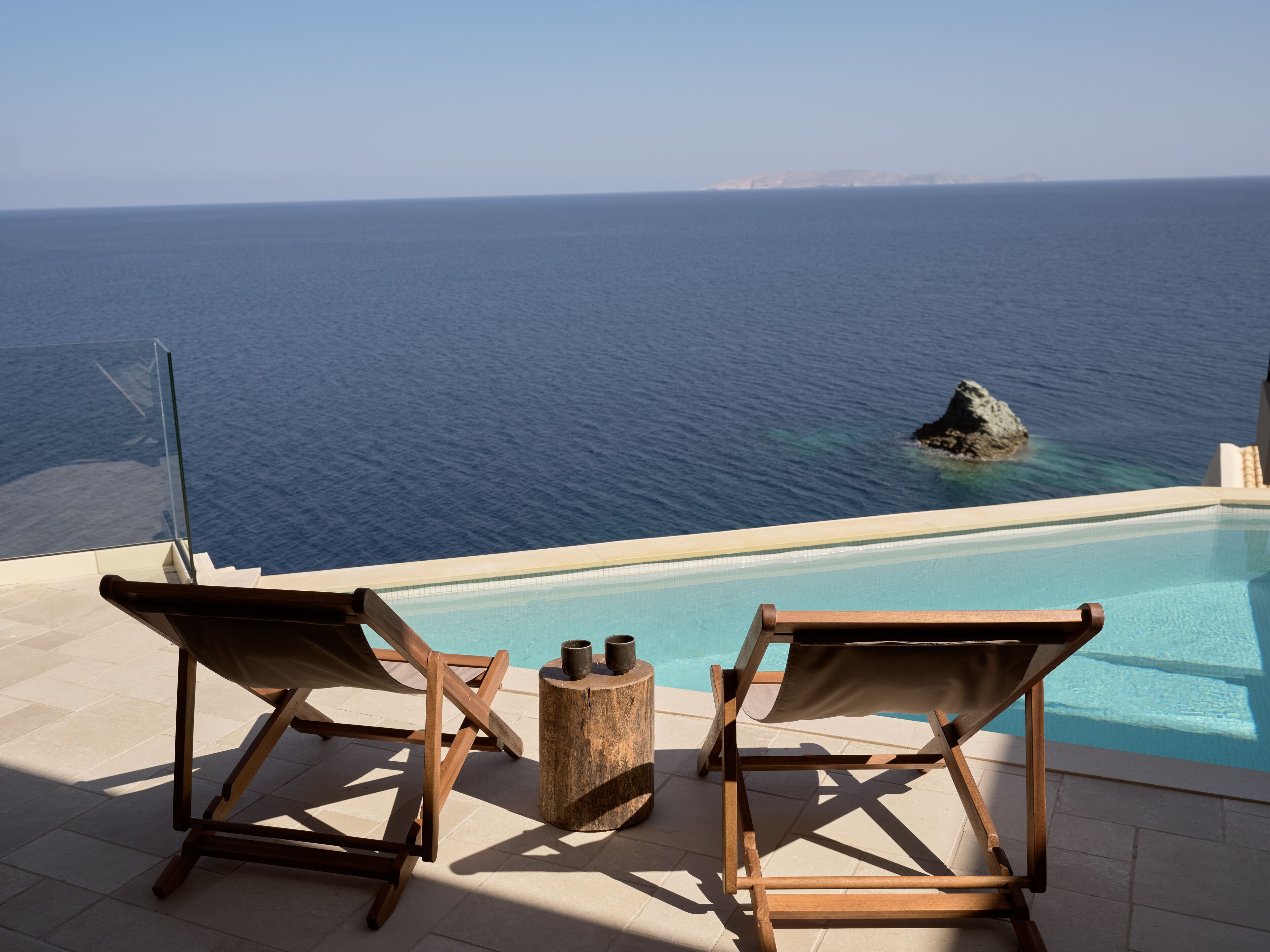 A poolside area with two wooden lounge chairs and a small table, overlooking a clear blue sea with a rocky outcrop.