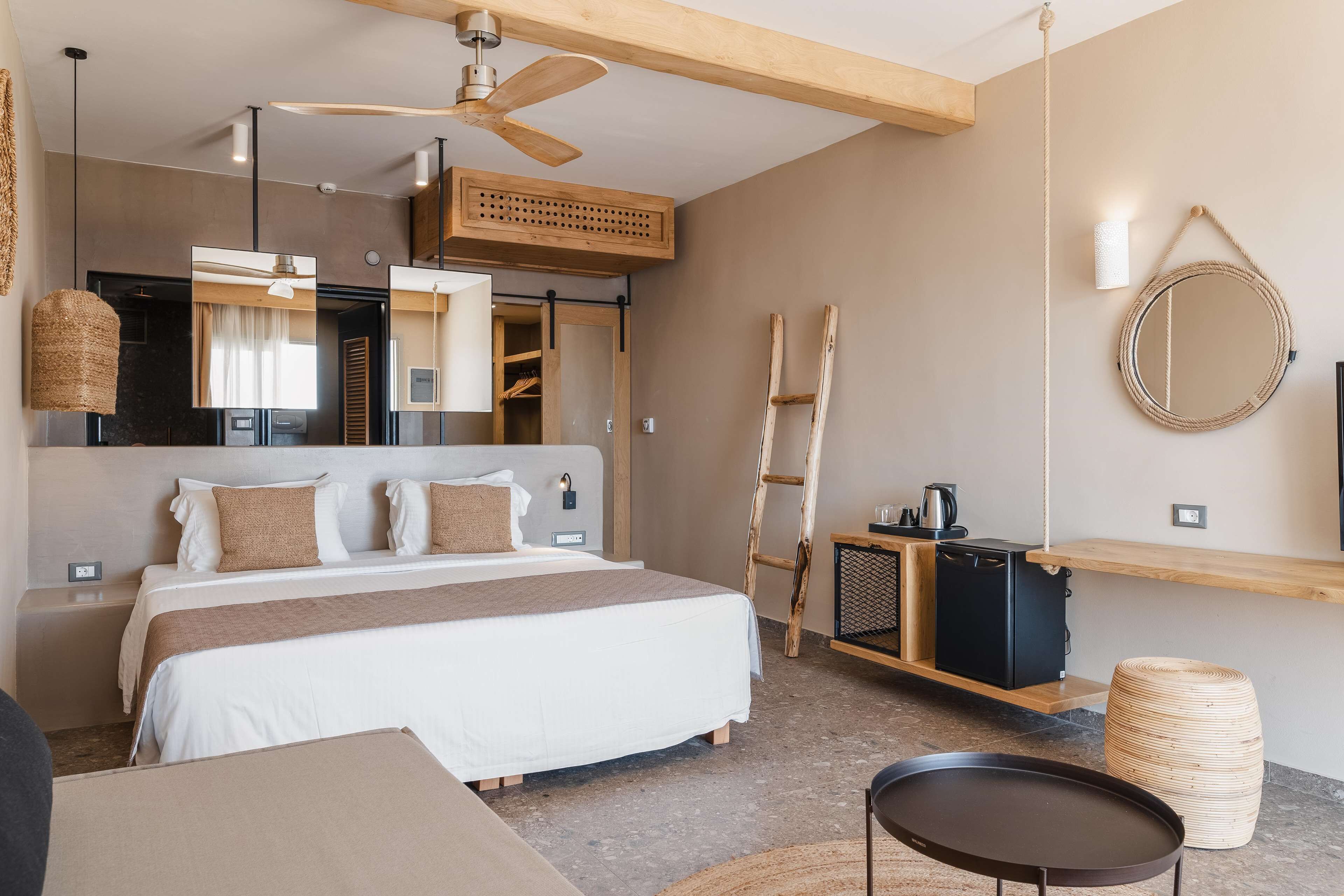 A modern hotel room with a large bed, a ceiling fan, a wooden ladder, a round mirror, and a small table.