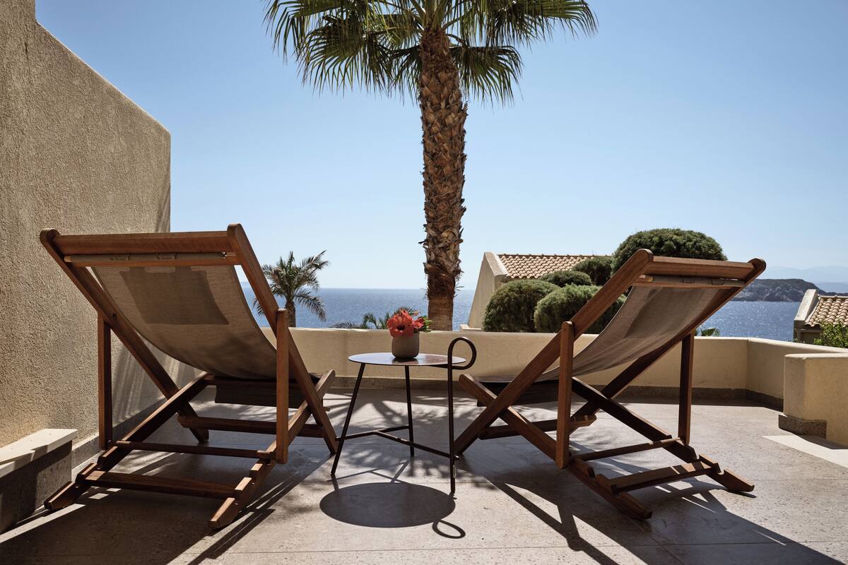 Two wooden lounge chairs, a small round table with a flower vase, and a view of the sea.
