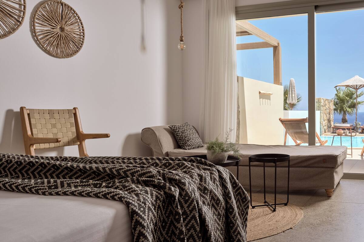 A modern hotel room with a bed, a sofa, a small table, and a view of a pool and palm trees.
