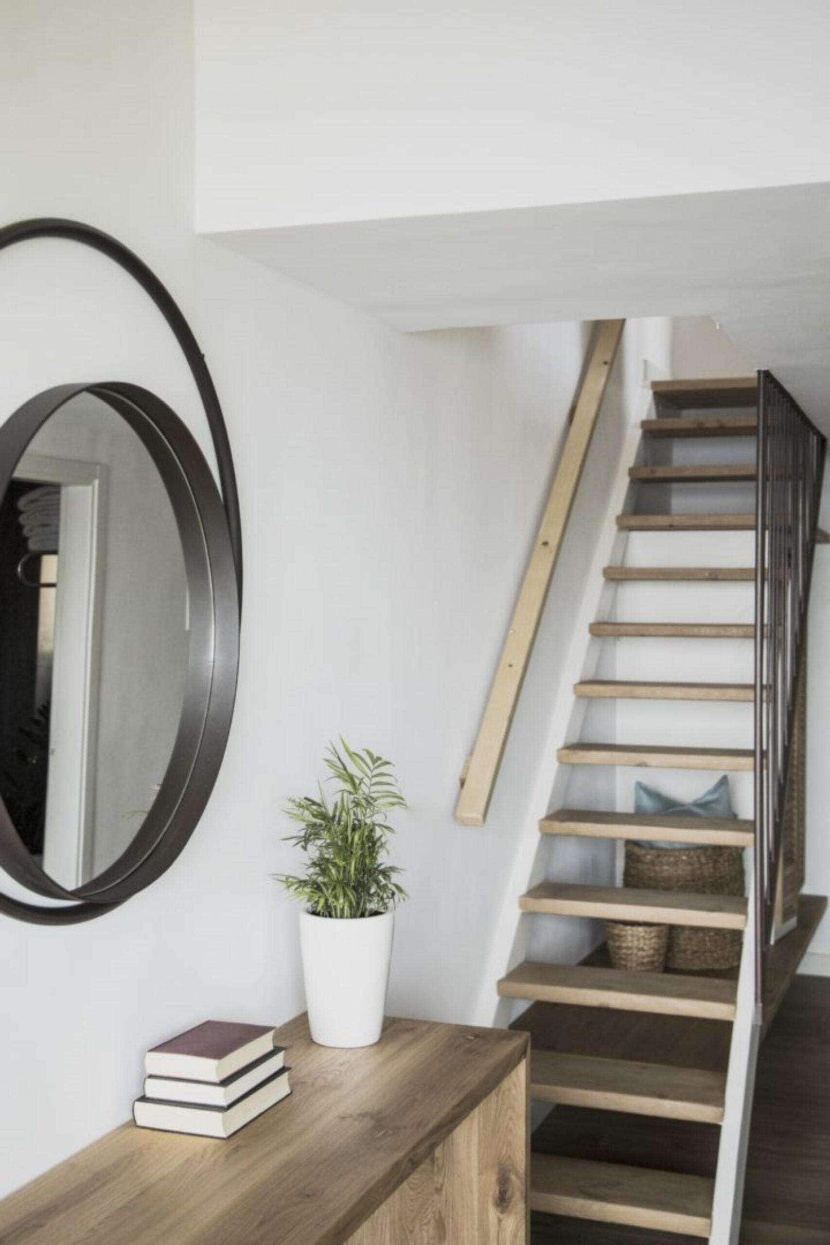 A staircase with wooden steps and a metal railing, a white wall, a round mirror, a potted plant, and a wooden shelf with books.