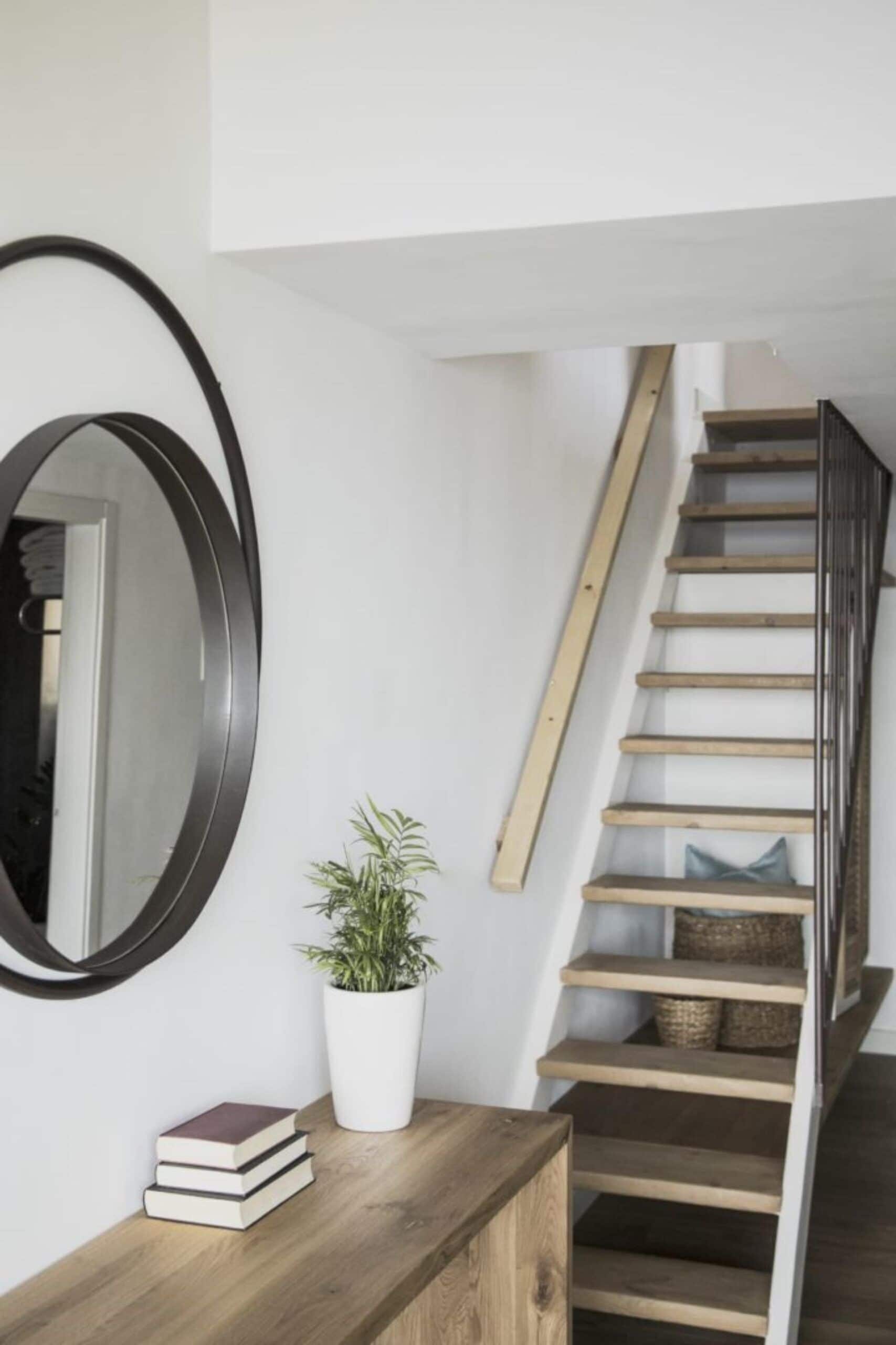 A staircase with wooden steps and a metal railing, a white wall, a round mirror, a potted plant, and a wooden console table with books.