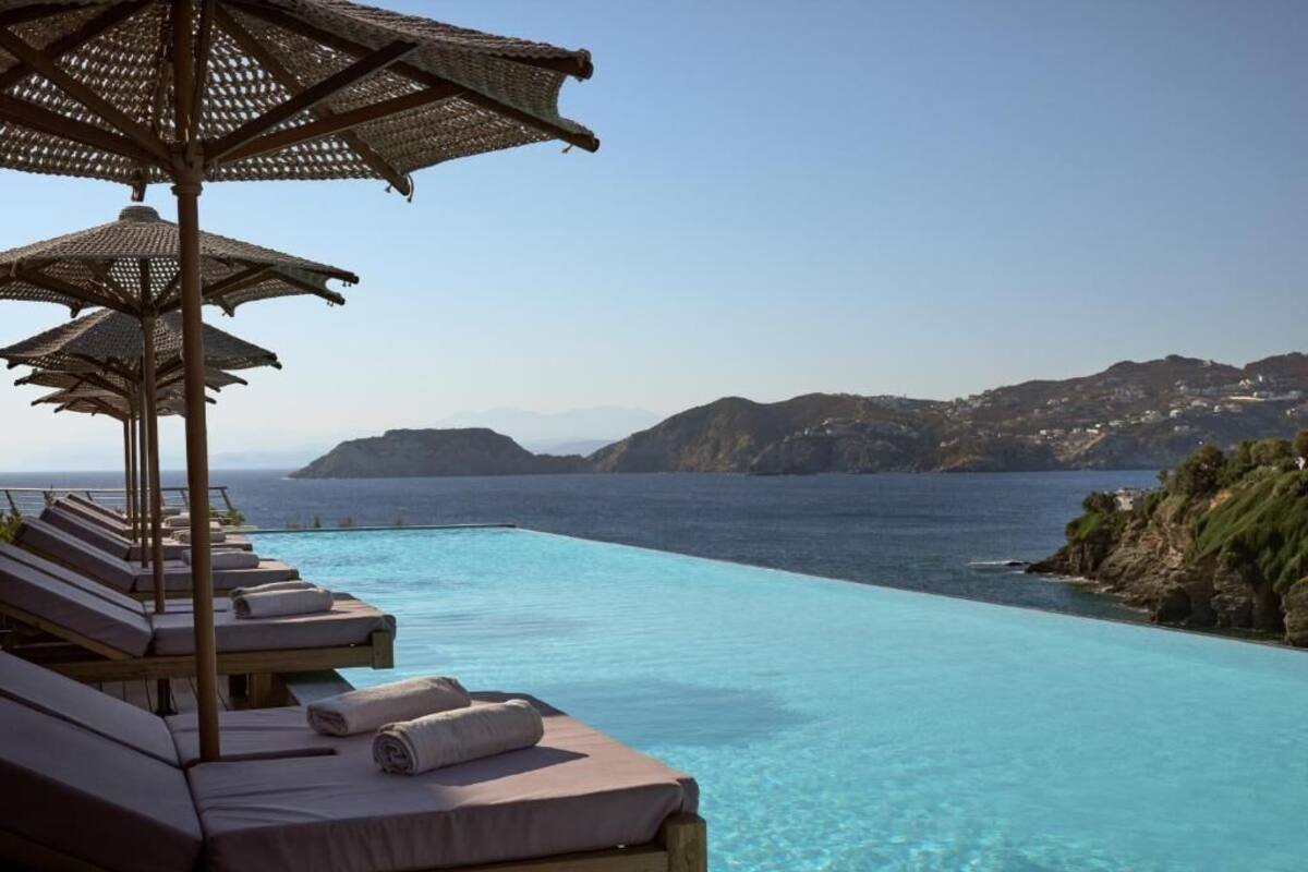 A poolside area with lounge chairs and umbrellas, overlooking a clear blue sea and distant hills.