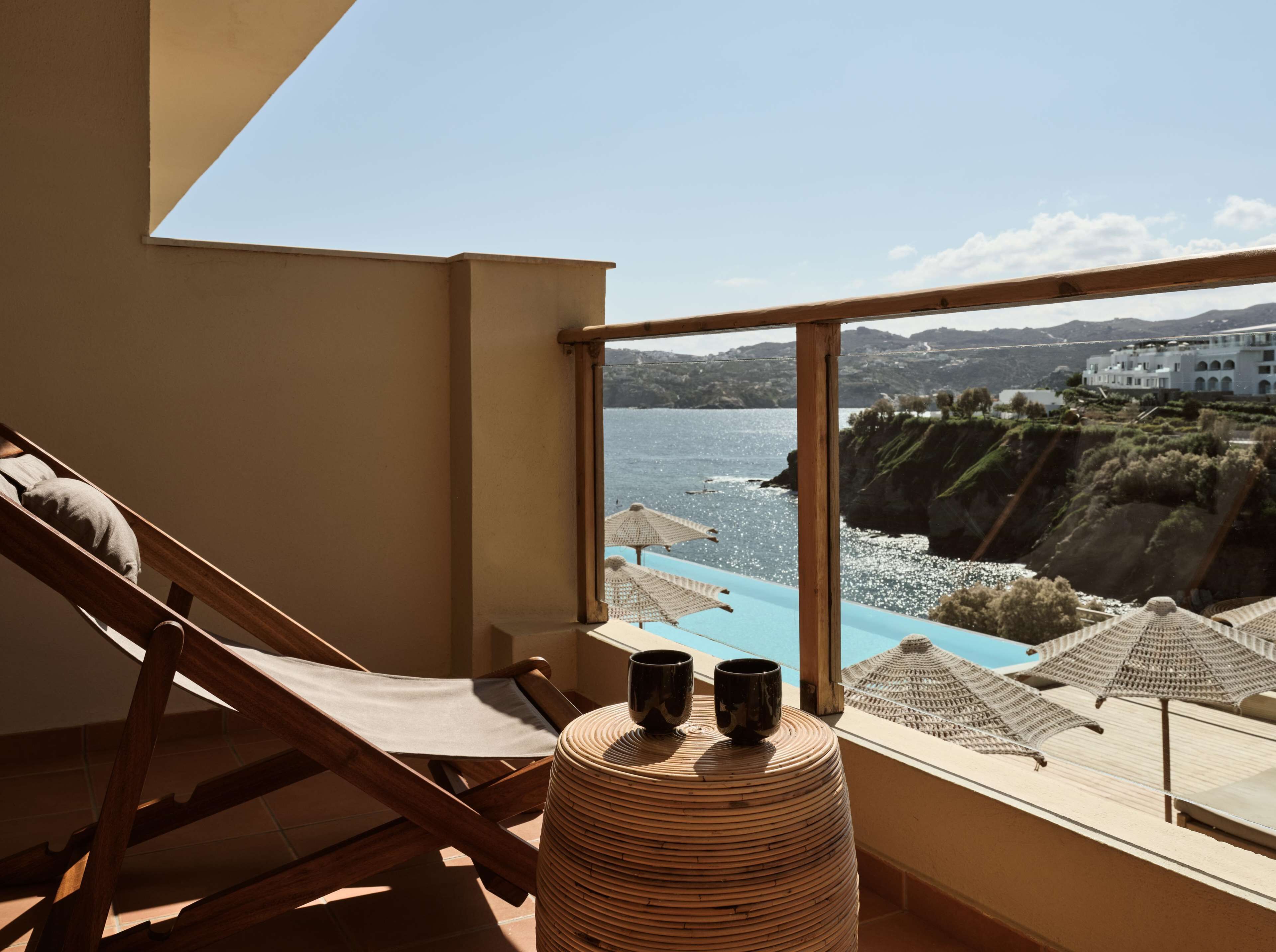 A balcony with a view of the sea, a wooden chair, and a woven stool with two cups.