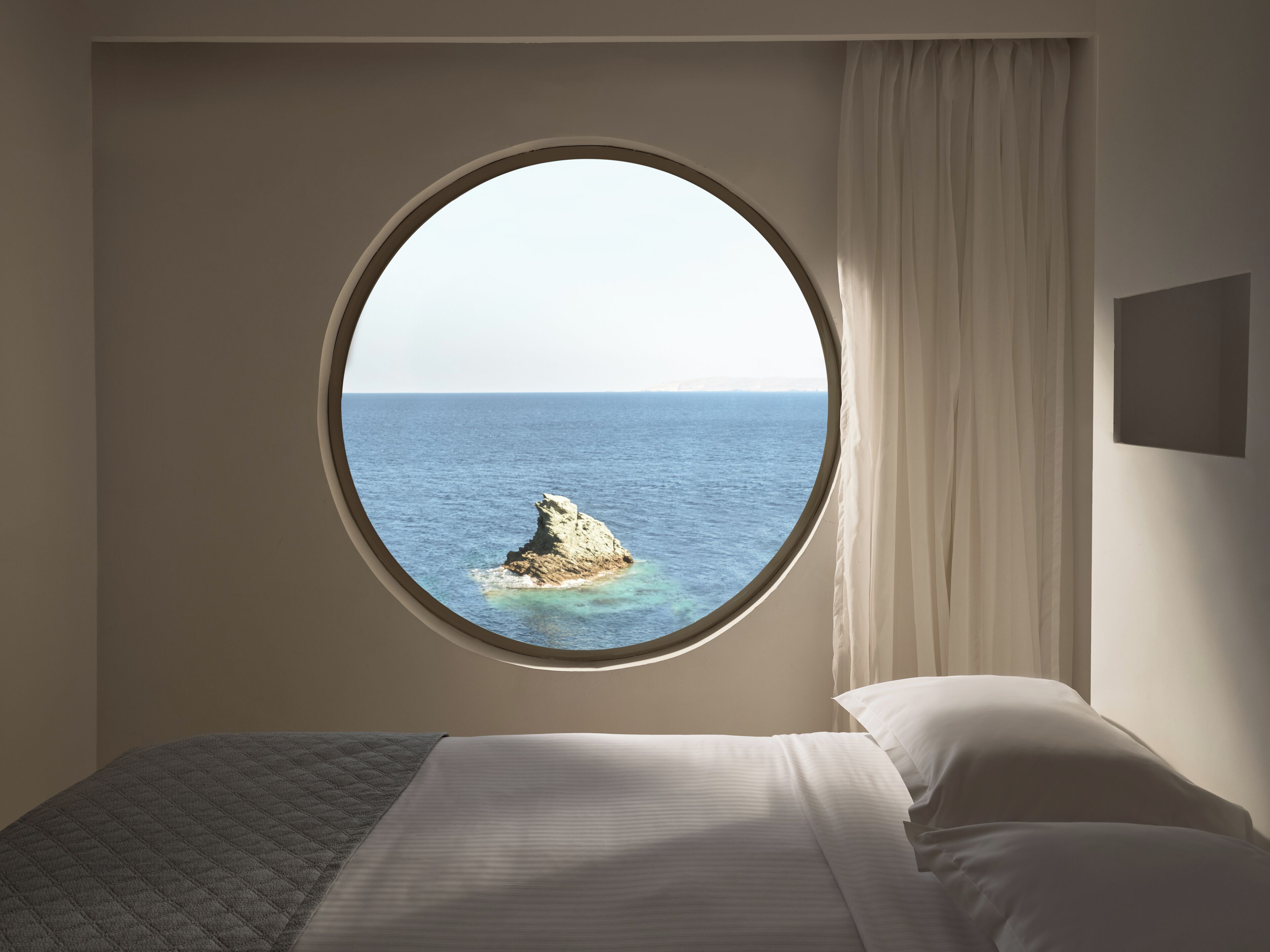 A bedroom with a round window overlooking the sea, a bed, and a curtain.
