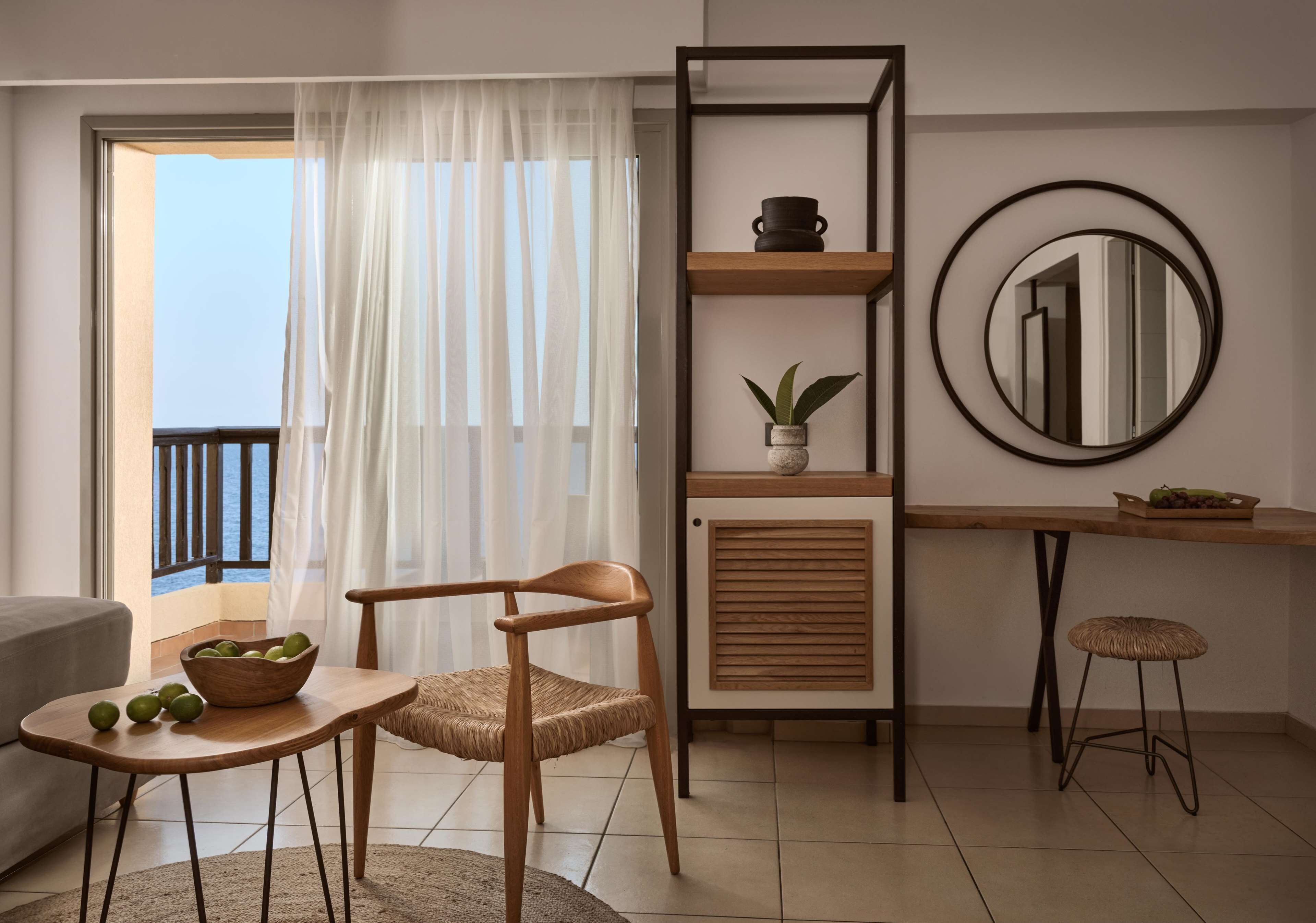 Un salon moderne avec une chaise en bois, une petite table sur laquelle se trouve un saladier de fruits, une étagère en bois ornée d’une plante, un miroir rond et une vue sur le balcon.