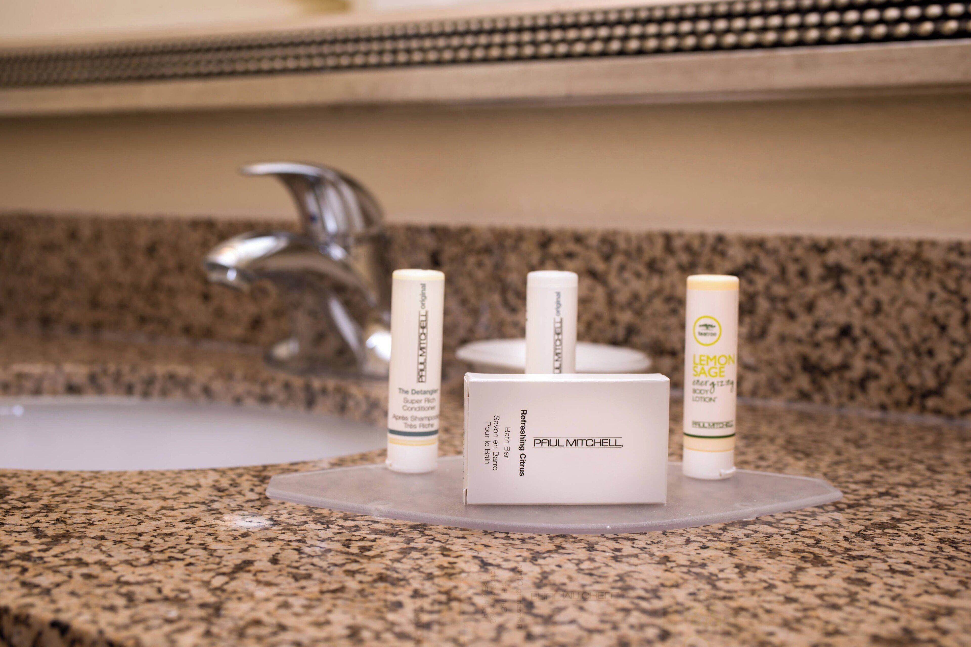 Una encimera de baño con cubierta de granito, un grifo plateado y tres botellas de artículos de aseo con las etiquetas "Paul Mitchell" y "Lemon Sage".