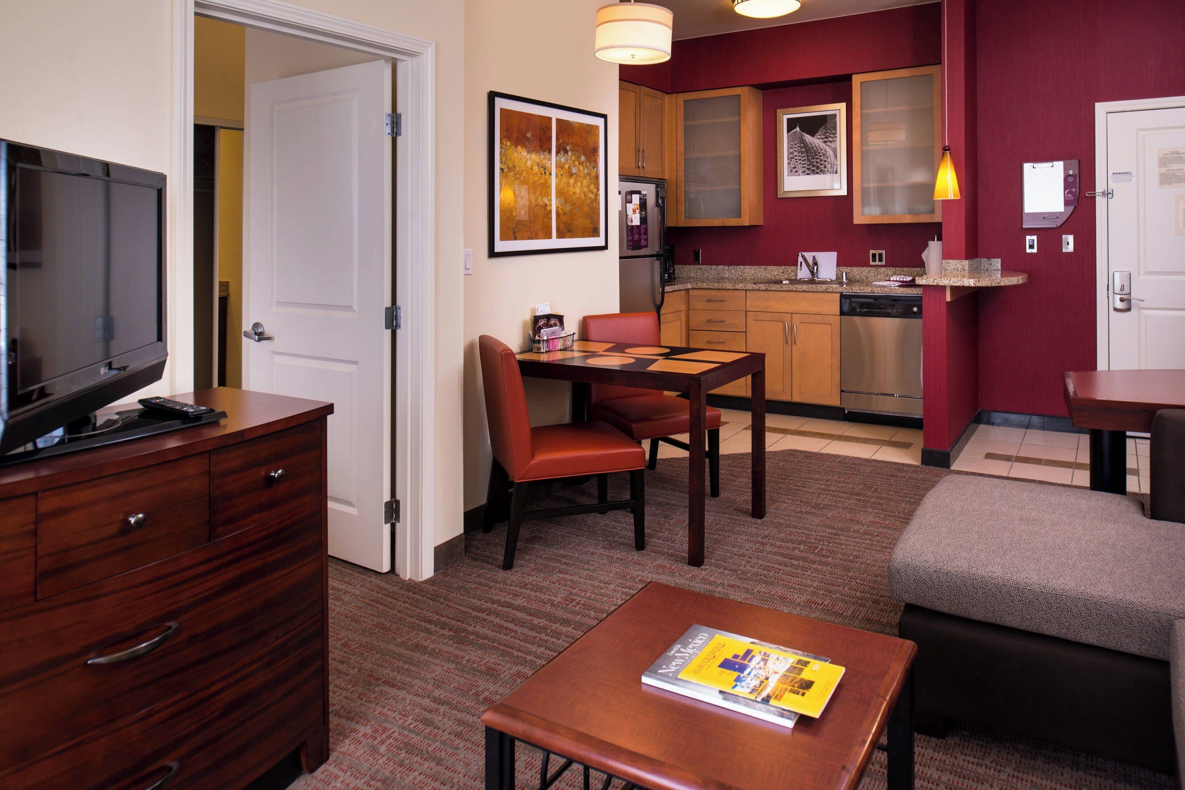 A hotel room with a TV, a dining table with chairs, a sofa, a small table with a magazine, a kitchen area with cabinets and a refrigerator, and a door leading to another room.