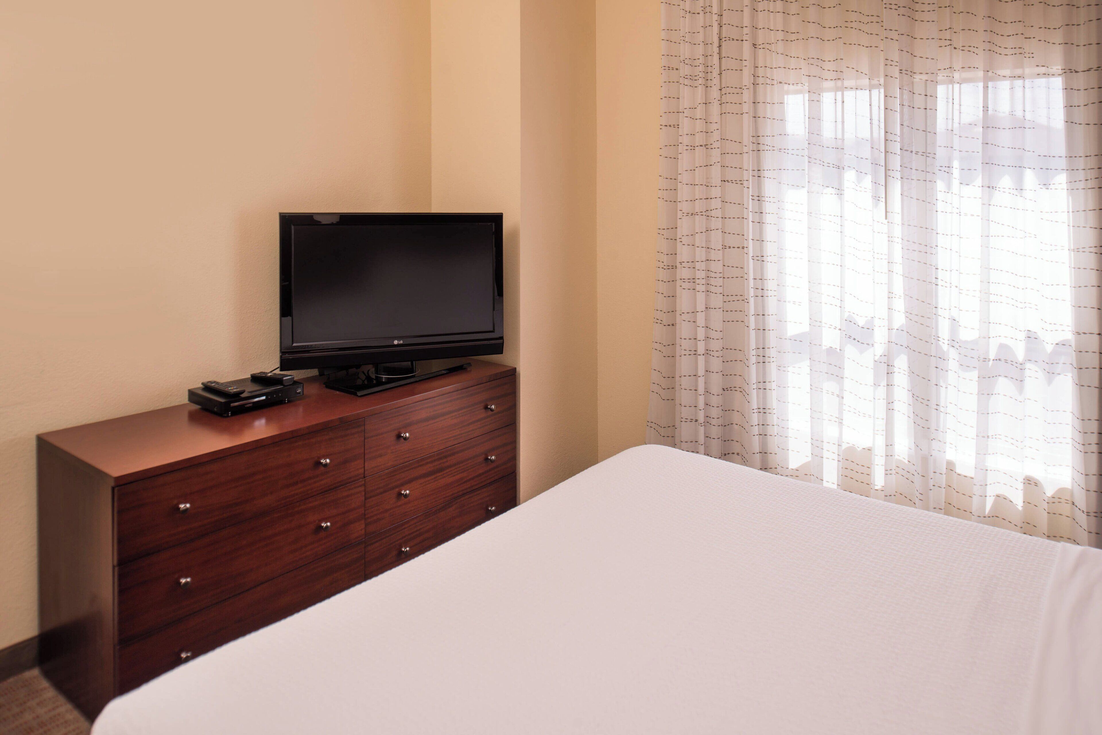 A hotel room with a bed, a wooden dresser with a flat-screen TV and remote control, and a window with sheer curtains.