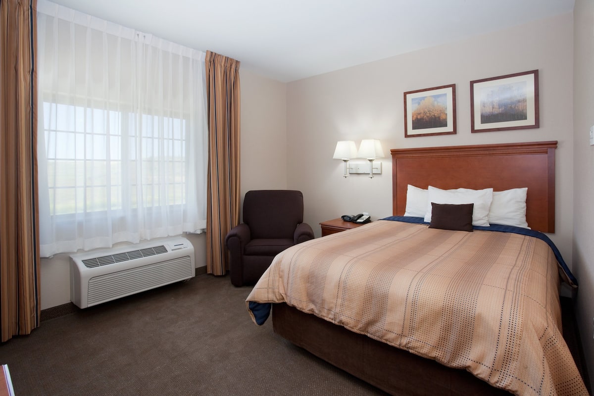 A hotel room with a bed, a chair, a bedside table with lamps, and a window with curtains.