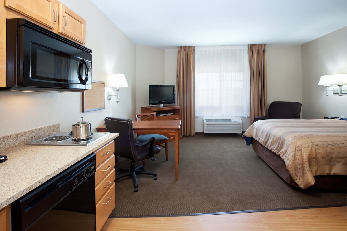 A hotel room with a kitchenette, a bed, a desk with a chair, a television, and a window with curtains.