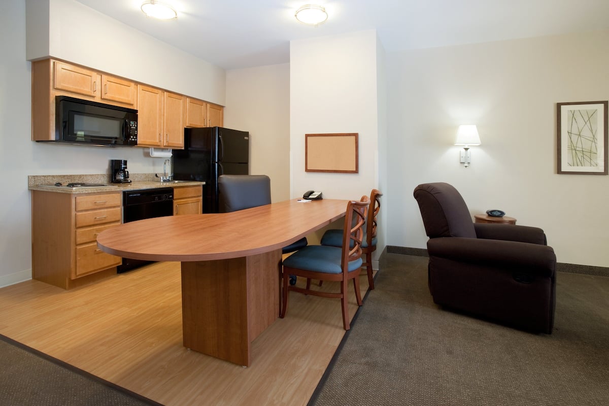 A compact kitchen with wooden cabinets, a microwave, and a refrigerator. There is a dining area with a wooden table and chairs, and a brown sofa. A lamp and framed artwork are on the wall.