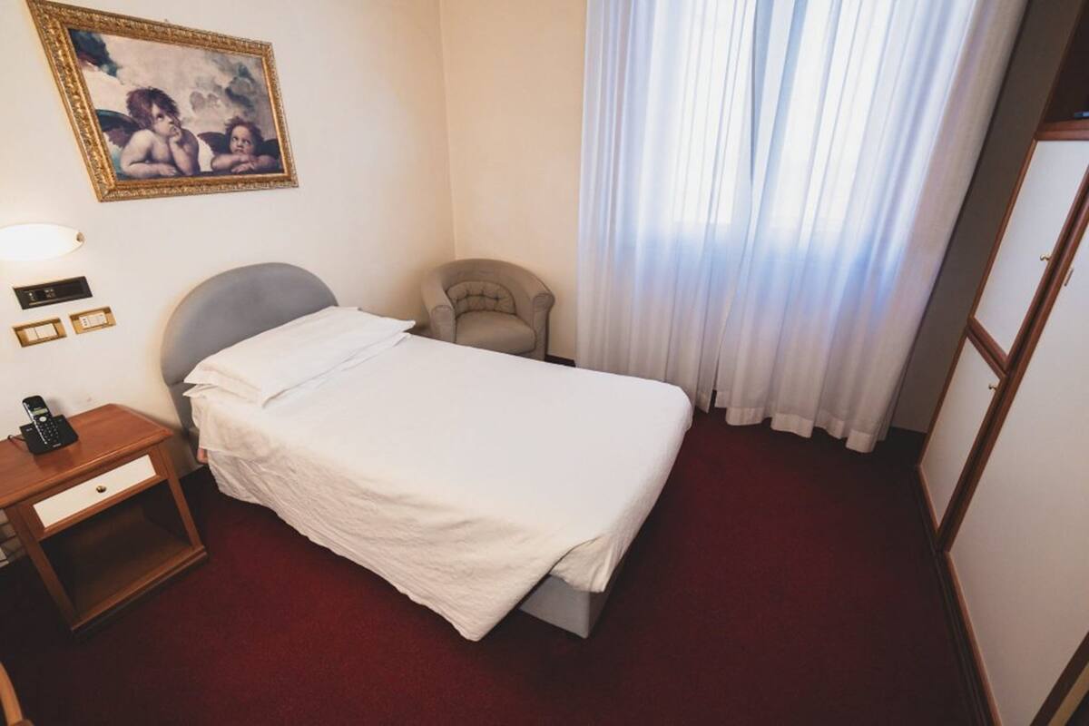 Een hotelkamer met een bed, een stoel, een nachtkastje met telefoon, een schilderij aan de muur en een raam met gordijnen.