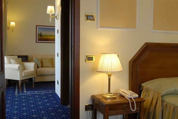 A hotel room with a bed, a bedside table with a lamp and phone, a chair, a sofa, and a patterned carpet.