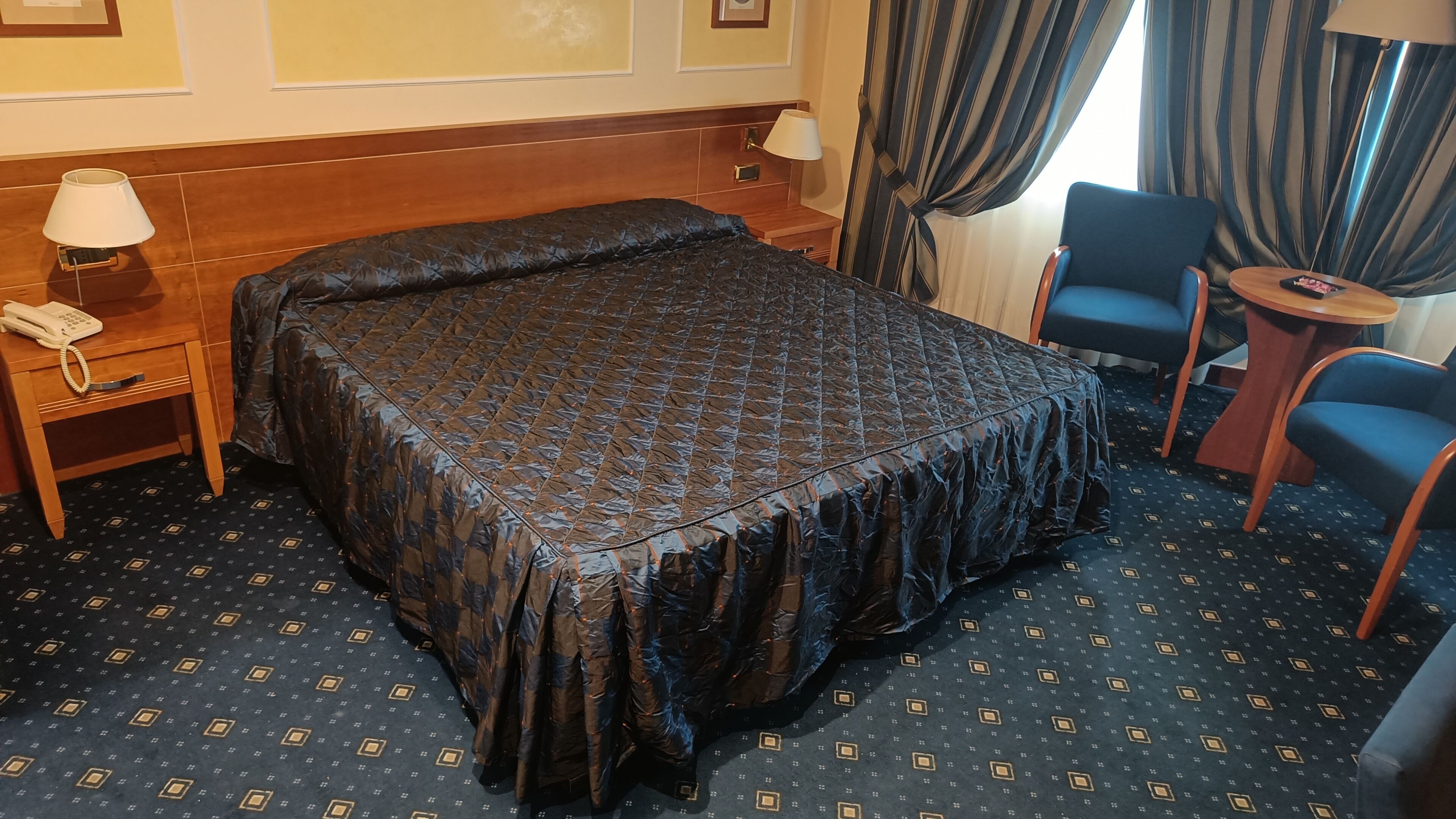 A hotel room with a large bed, a bedside table with a lamp, a chair, and a small table.