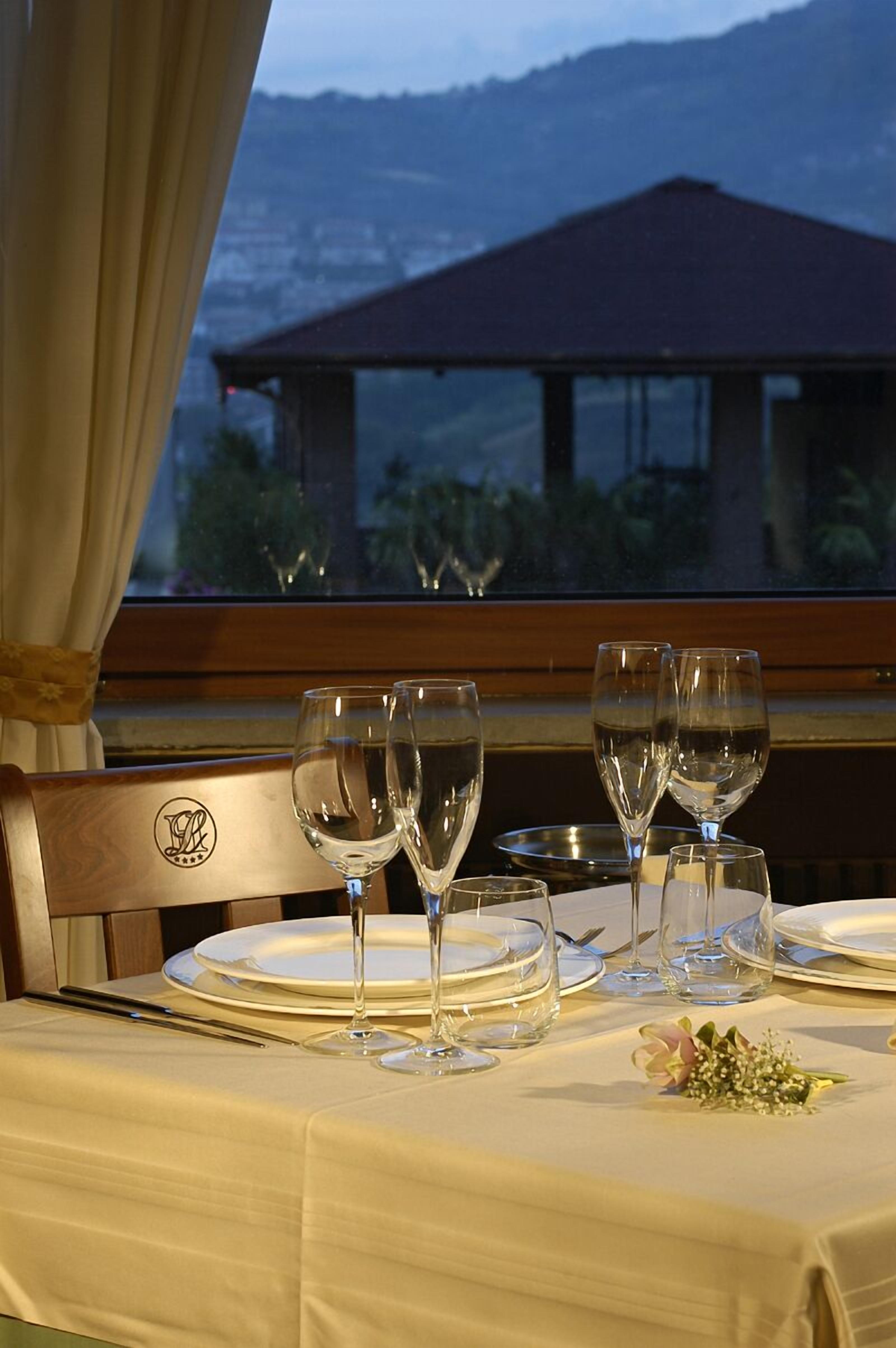 Mesa de comedor dispuesta con copas de vino, platos y cubiertos, con vista a una montaña y un edificio al fondo.