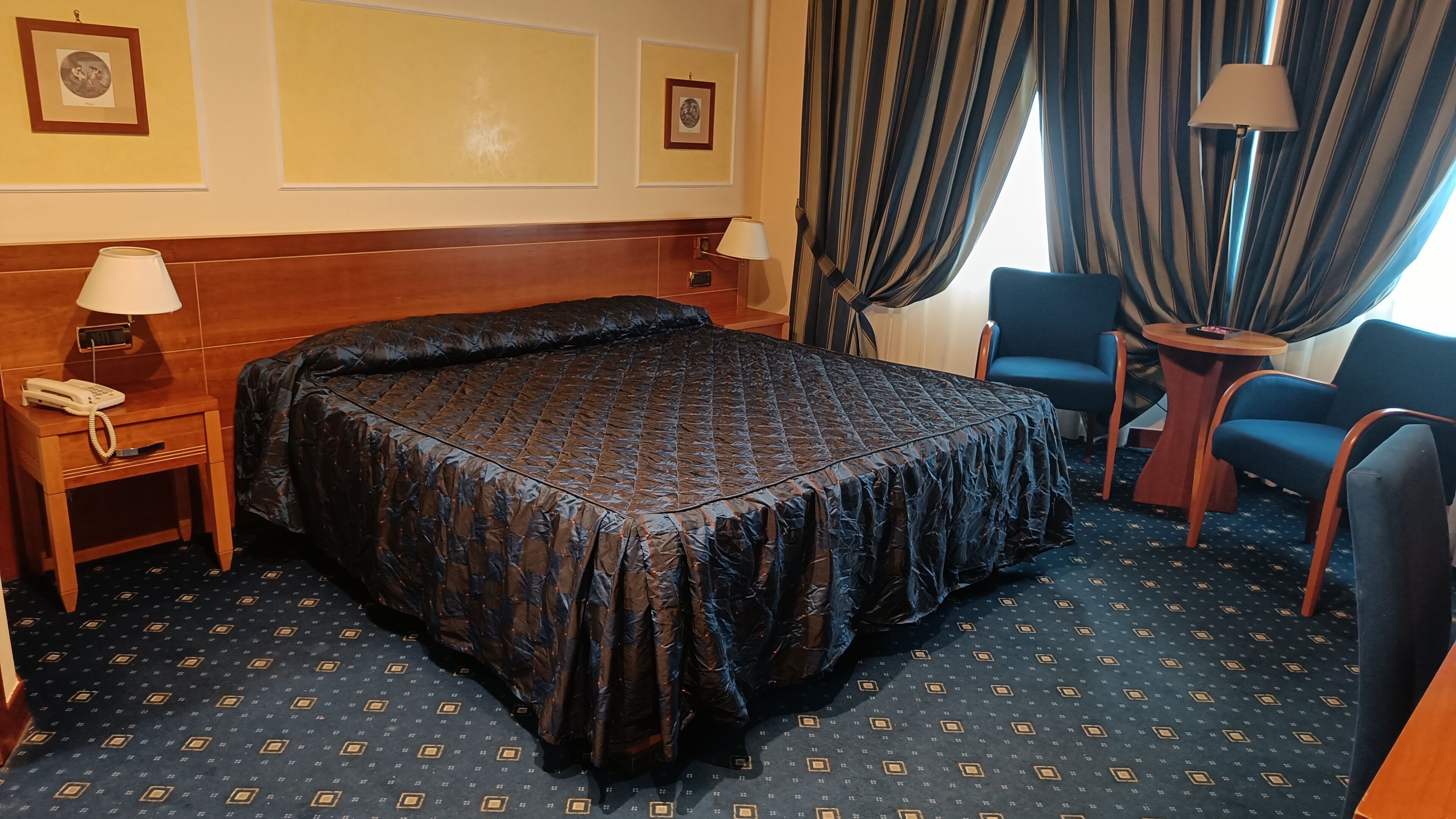 Habitación de hotel con una cama grande, una mesita de noche con lámpara, una silla y una pequeña mesa con lámpara.