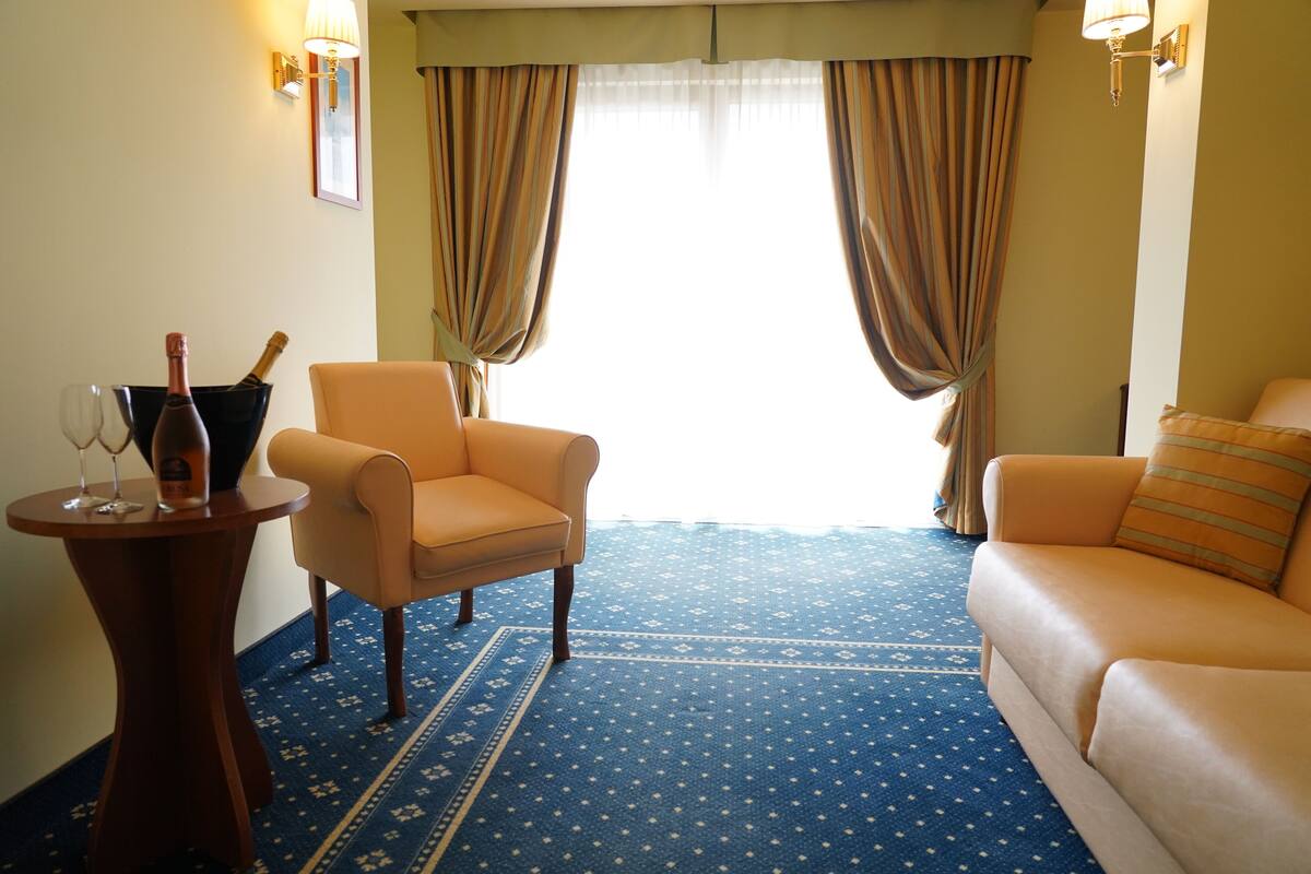 Una habitación con alfombra azul estampada, un sofá color crema, una mesita redonda con una botella y vasos, y cortinas en las ventanas.