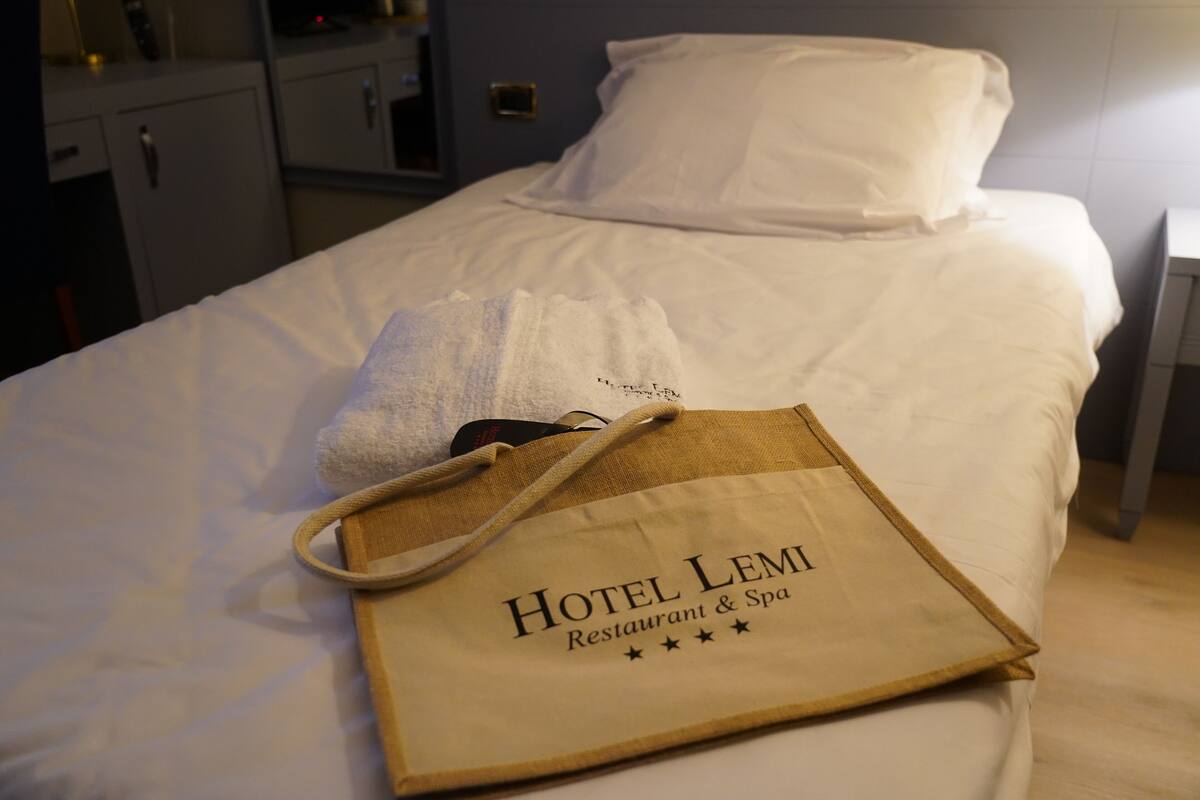 Una cama bien hecha con una almohada blanca y una toalla, un bolso de madera con el texto 'Hotel Lemi Restaurant & Spa' y una pequeña mesita de noche.