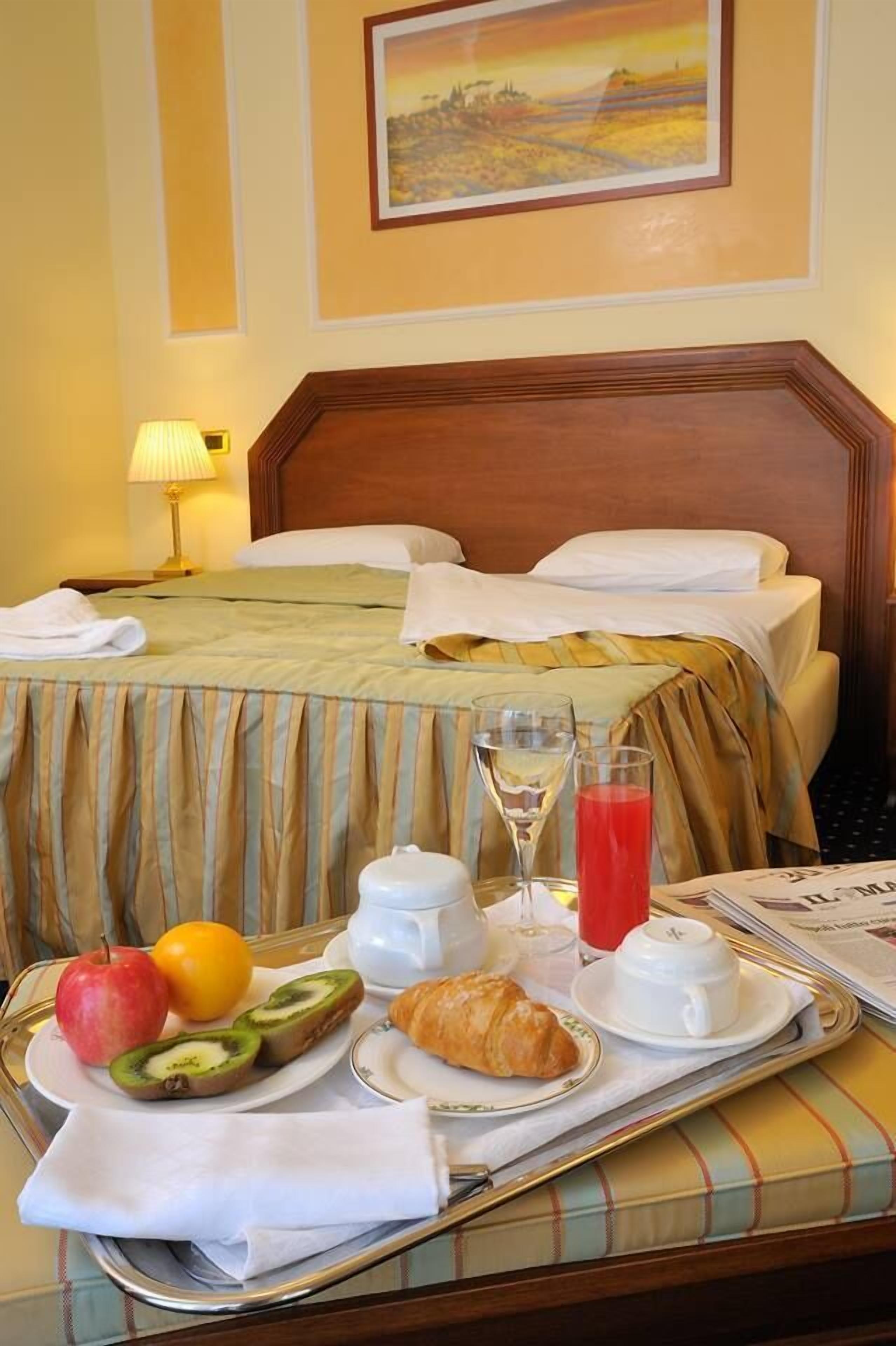 Habitación de hotel con una cama bien hecha, una bandeja con alimentos para el desayuno, incluyendo frutas, un croissant y bebidas, y un cuadro en la pared.