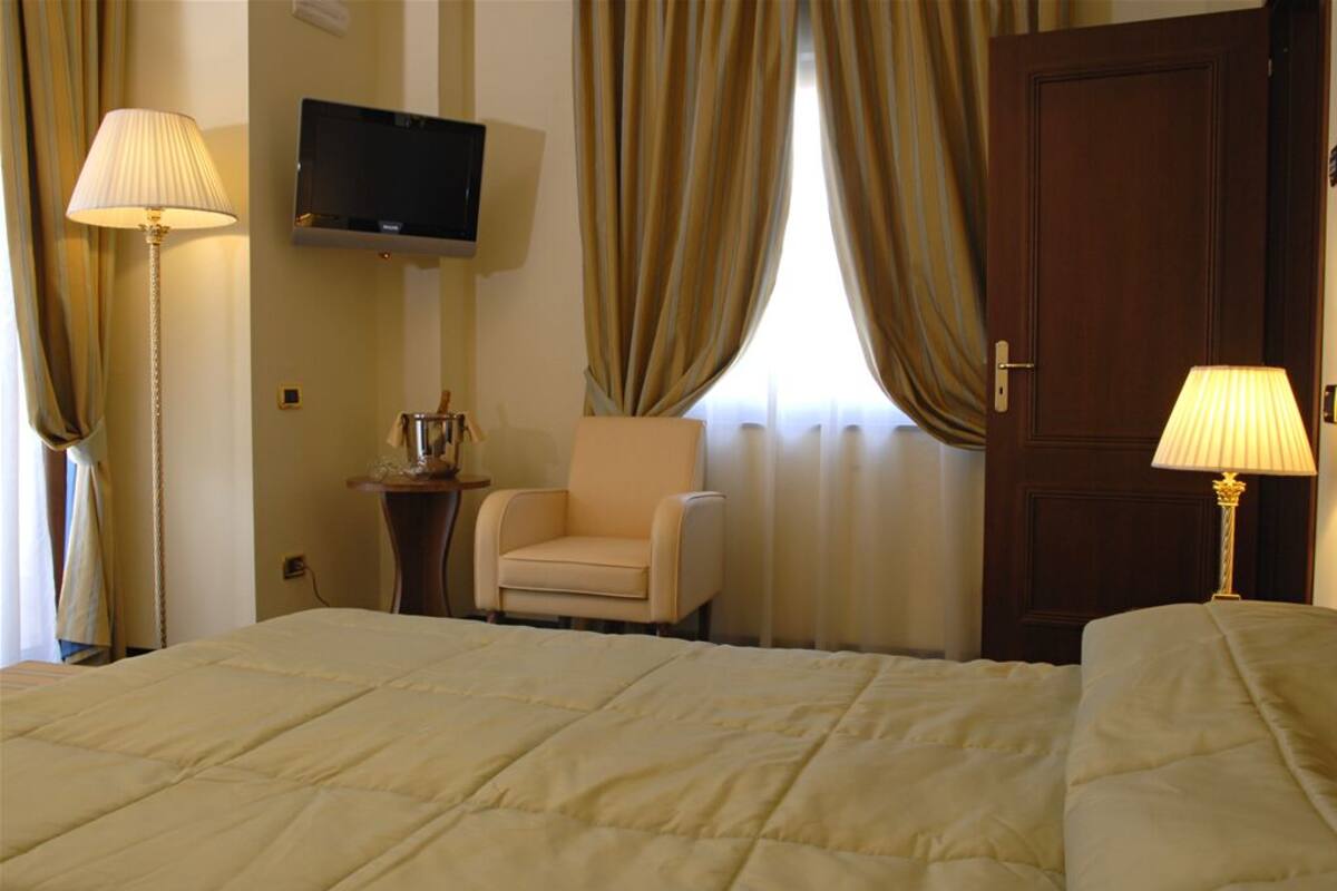 Una habitación de hotel con una cama, dos lámparas de noche, un televisor, un sillón y una puerta.