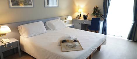 A hotel room with a large bed, bedside tables with lamps, a desk, and a chair. There are framed pictures on the wall and a window with curtains.