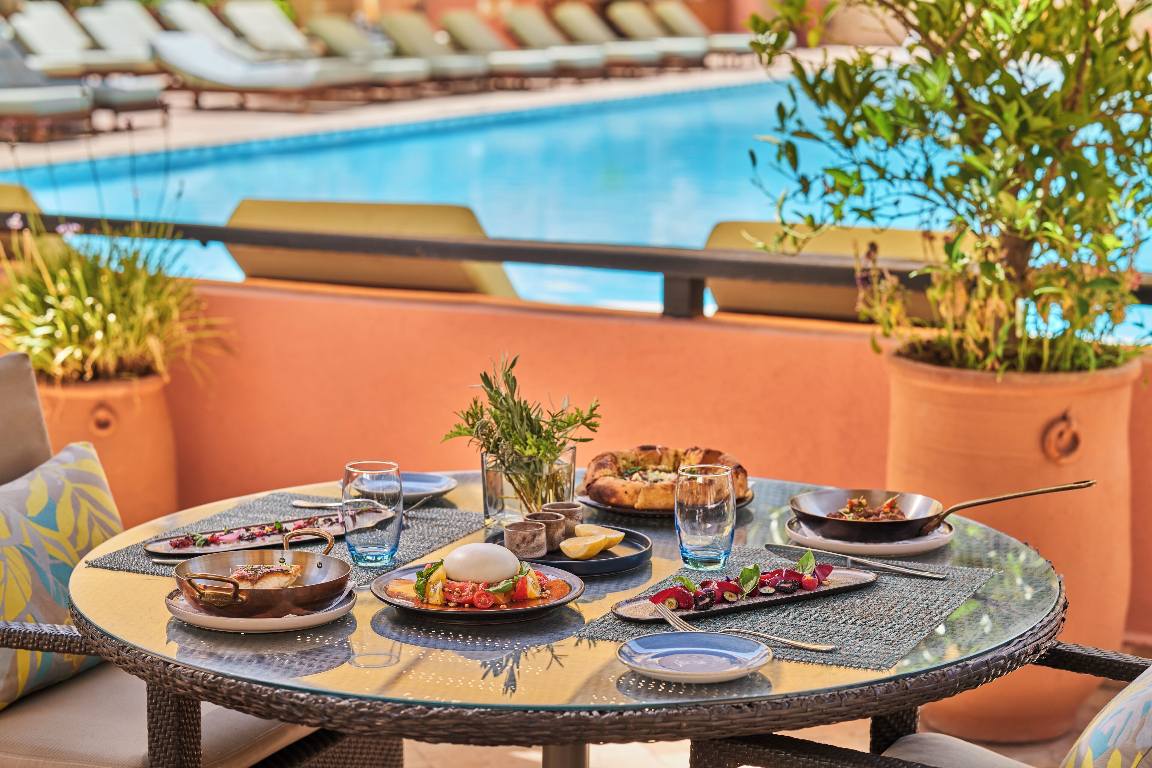 Ett runt glasbord med olika rätter, ett glas vatten och en pizza på en terrass med en pool i bakgrunden.