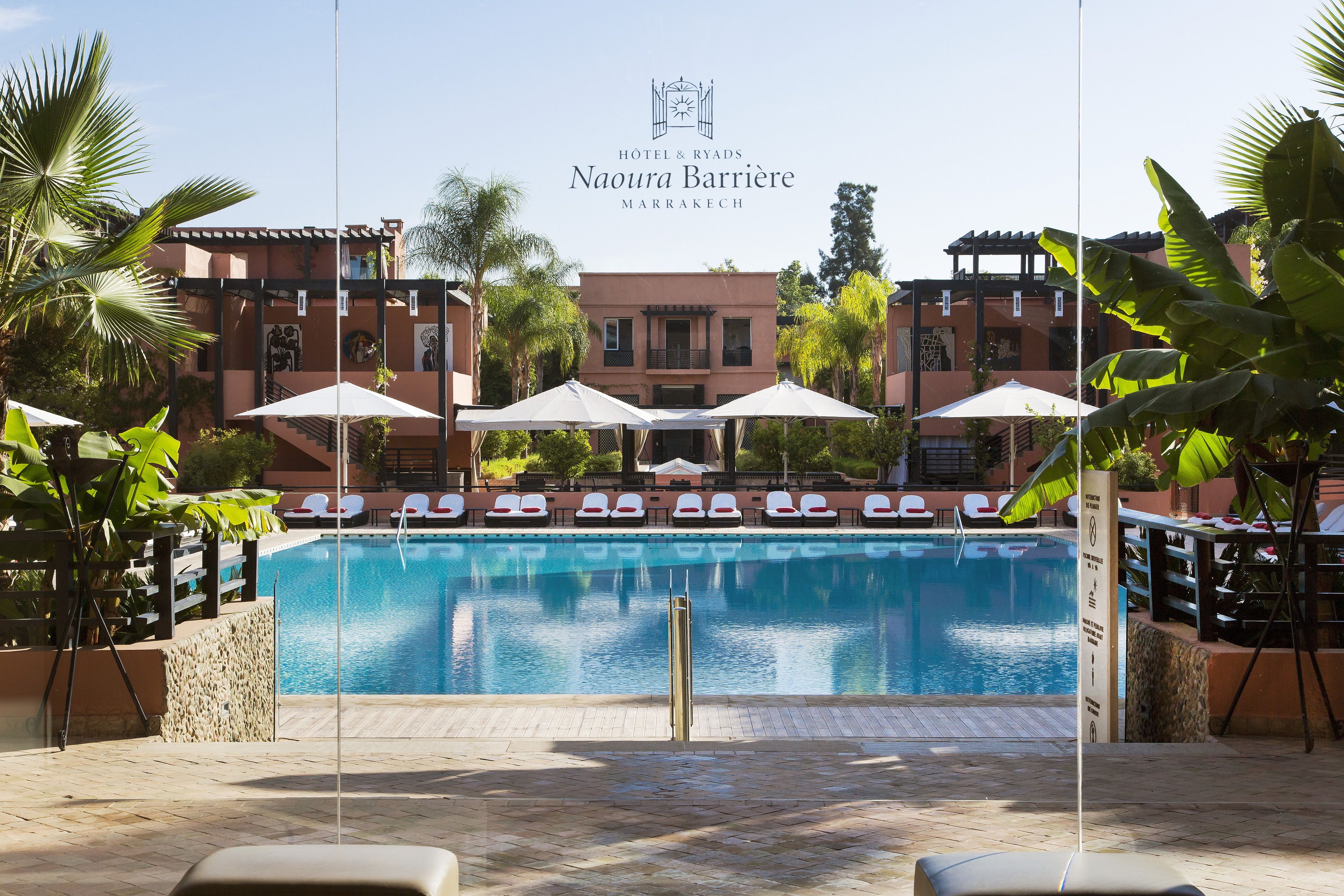 Piscina com espreguiçadeiras e guarda-sóis, rodeada por palmeiras e um edifício com o letreiro "Hotel & Ryads Naoura Barrie Marrakech".