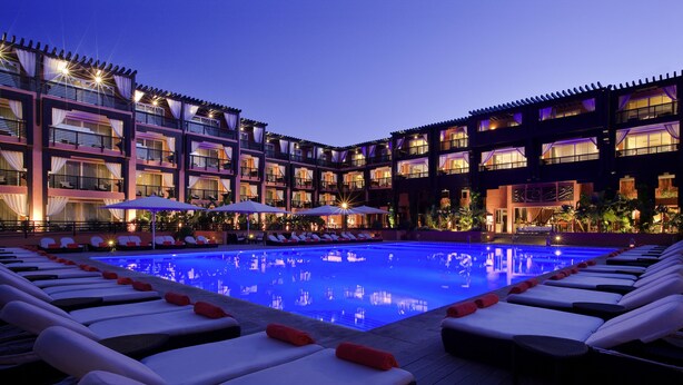 A hotel with a poolside area featuring sun loungers and umbrellas, a multi-story building with balconies, and a well-lit facade.
